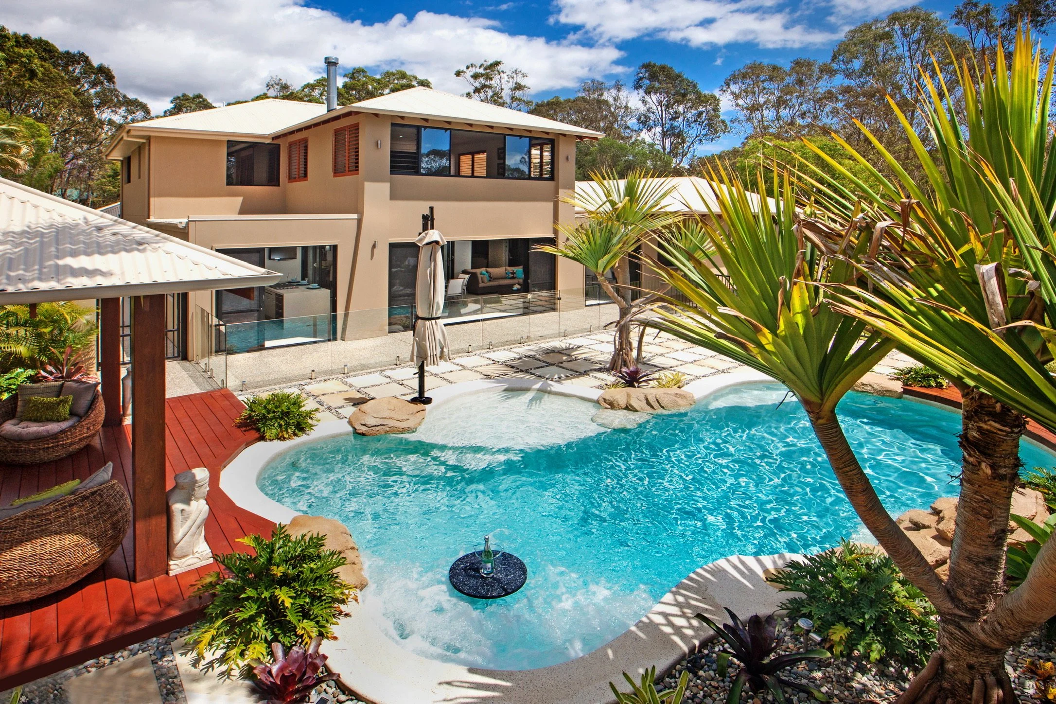 A modern house with a backyard featuring a small pool surrounded by plants, outdoor seating, and a glass barrier with trees and a blue sky in the background.