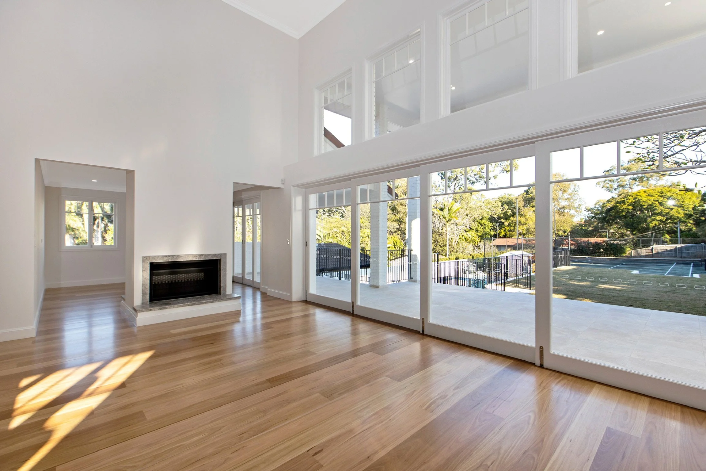 Bright living room with large glass windows and doors overlooking a backyard with trees and a tennis court, hardwood flooring, and a white ceiling and walls.