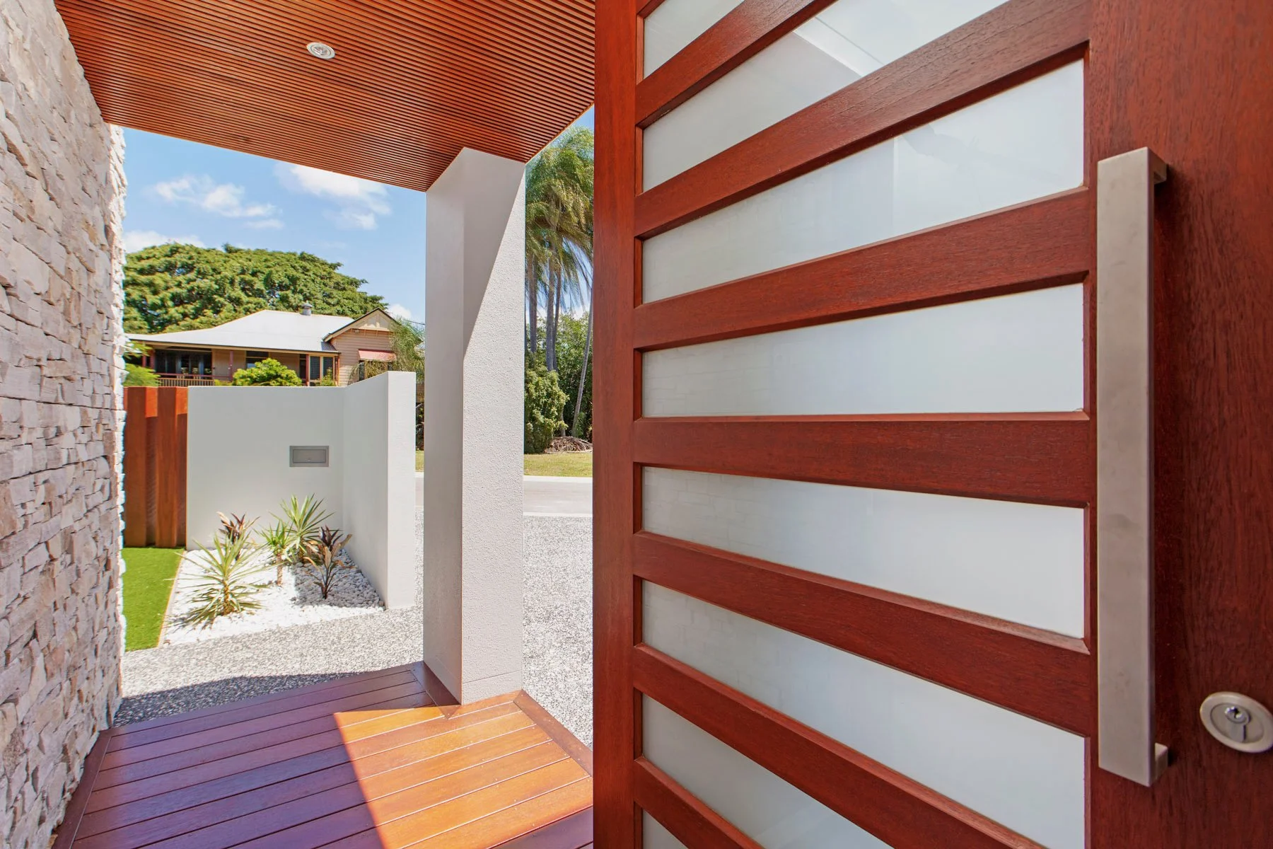 View of a modern house entrance with a wooden gate and stone walls, featuring a small garden with plants and white gravel, under a blue sky with clouds.