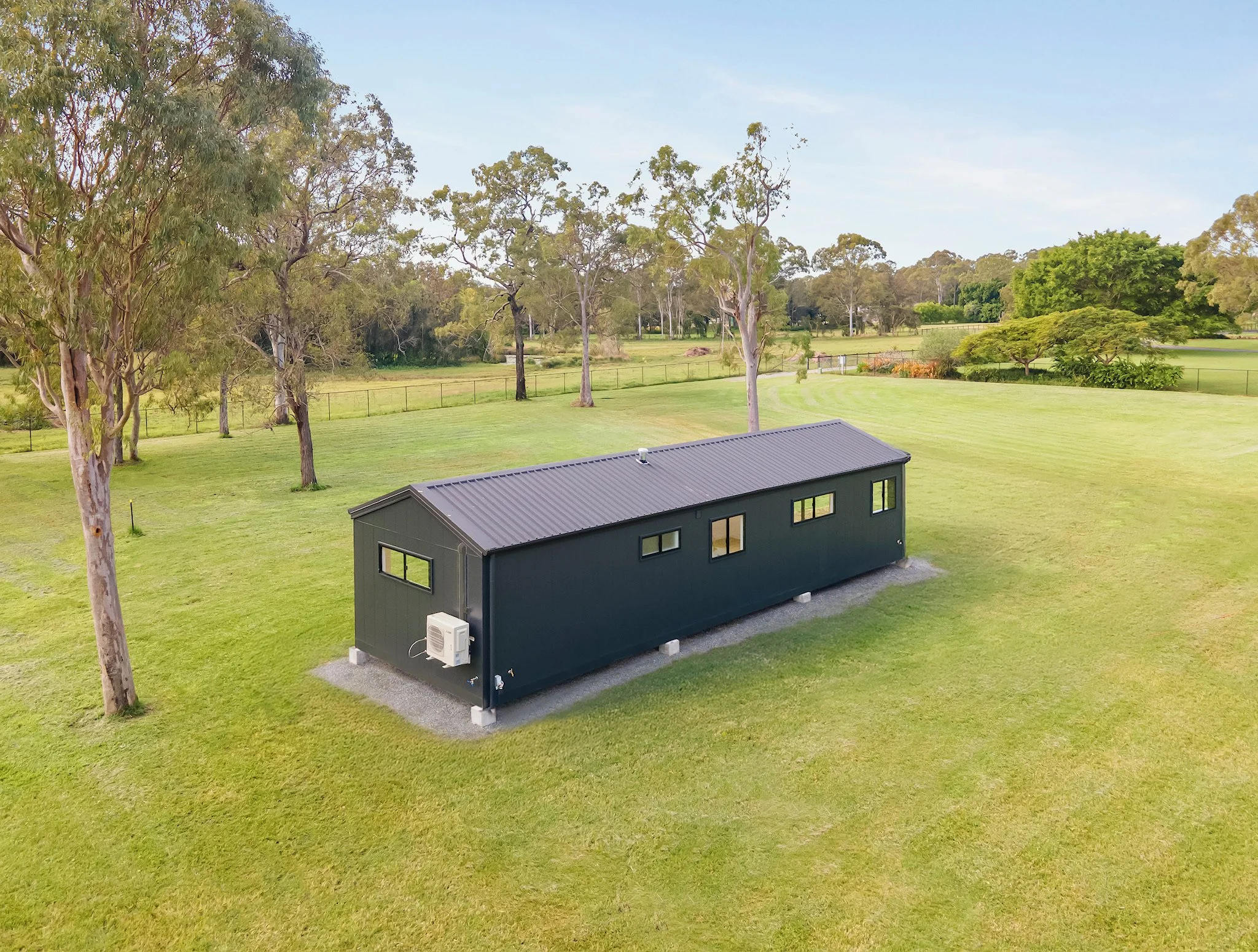 A modern, dark gray tiny house with a metal roof situated on a grassy lawn surrounded by trees and open land.