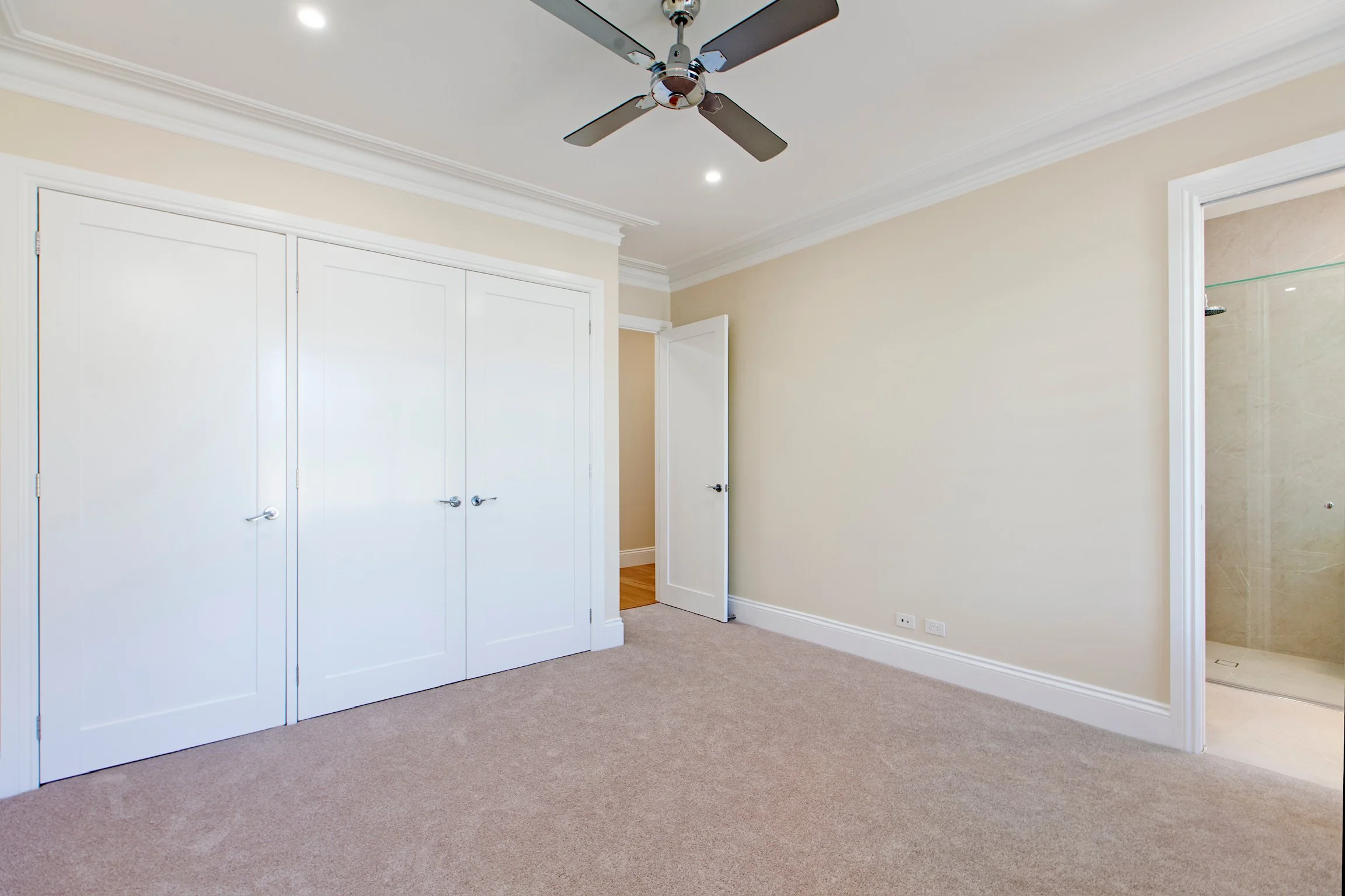 An empty bedroom with beige carpet, white walls, a white double closet door, a partially open door, and an open bathroom door with a glass shower. A ceiling fan with four blades and modern lighting is mounted on the ceiling.