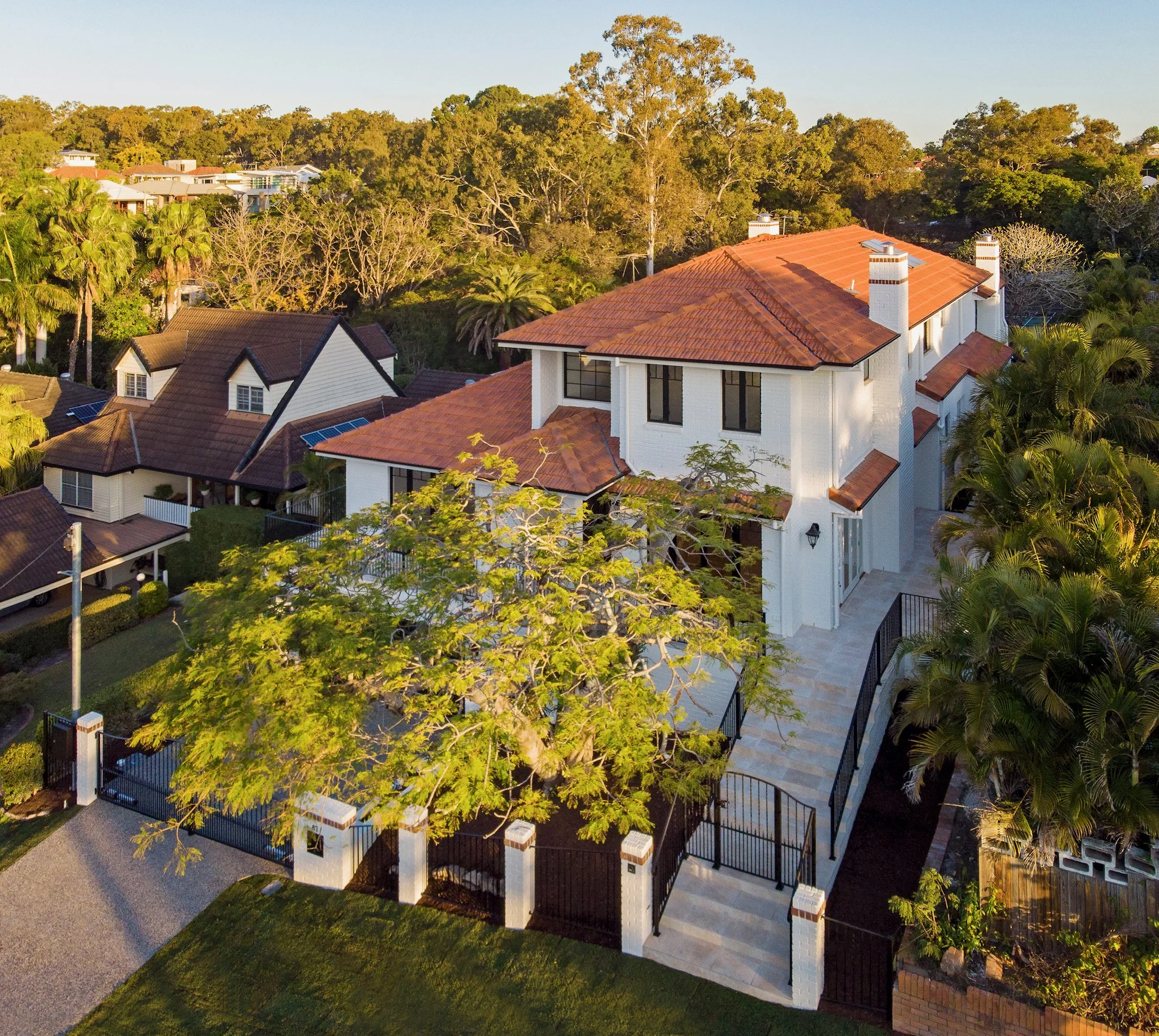 Aerial view of suburban houses with red-tile roofs surrounded by green trees under a clear blue sky during daylight.