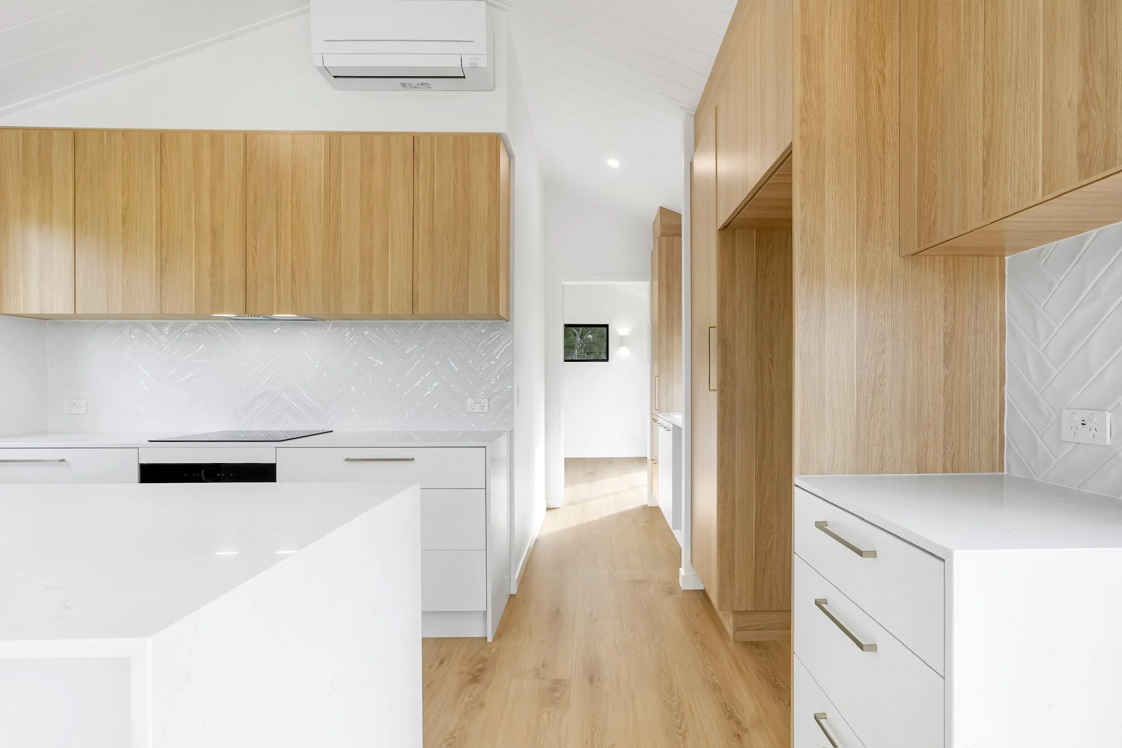 Modern kitchen with wooden cabinetry, white lower cabinets, and white tiled backsplash, with visible kitchen appliances and a window in the background.