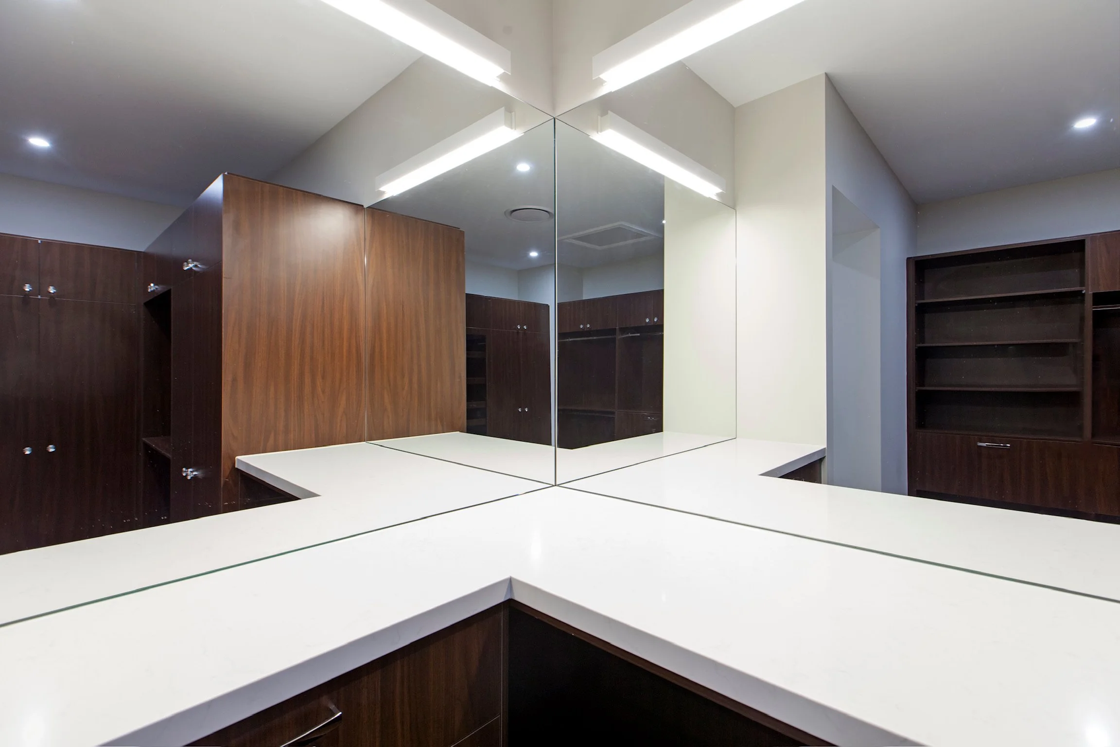 A modern bathroom with white countertops, methacrylate glass mirrors, and dark wood cabinets. The ceiling has integrated lighting fixtures, and the walls are painted white.
