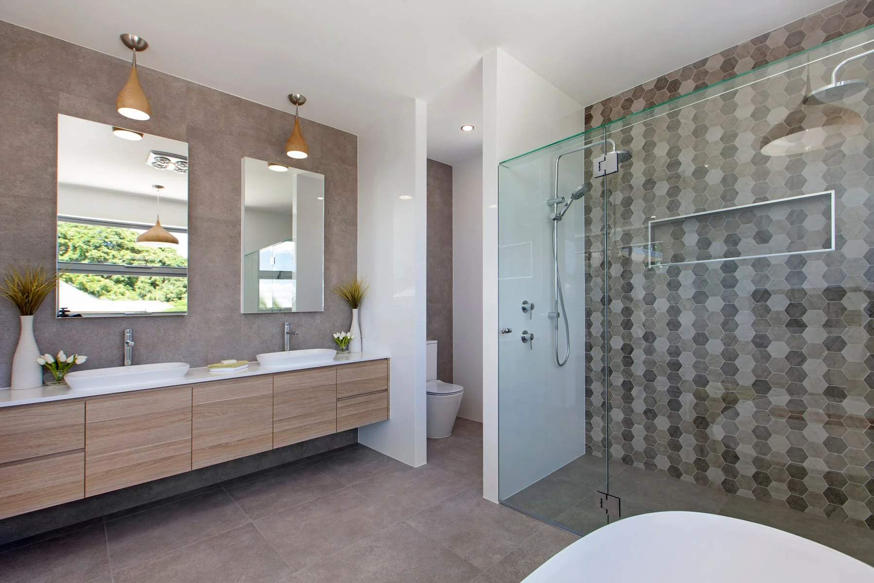 Modern bathroom with dual sinks, large mirrors, a walk-in shower with hexagonal tile walls, and a toilet. Decor includes vases with flowers and plants, with natural light coming through a window.