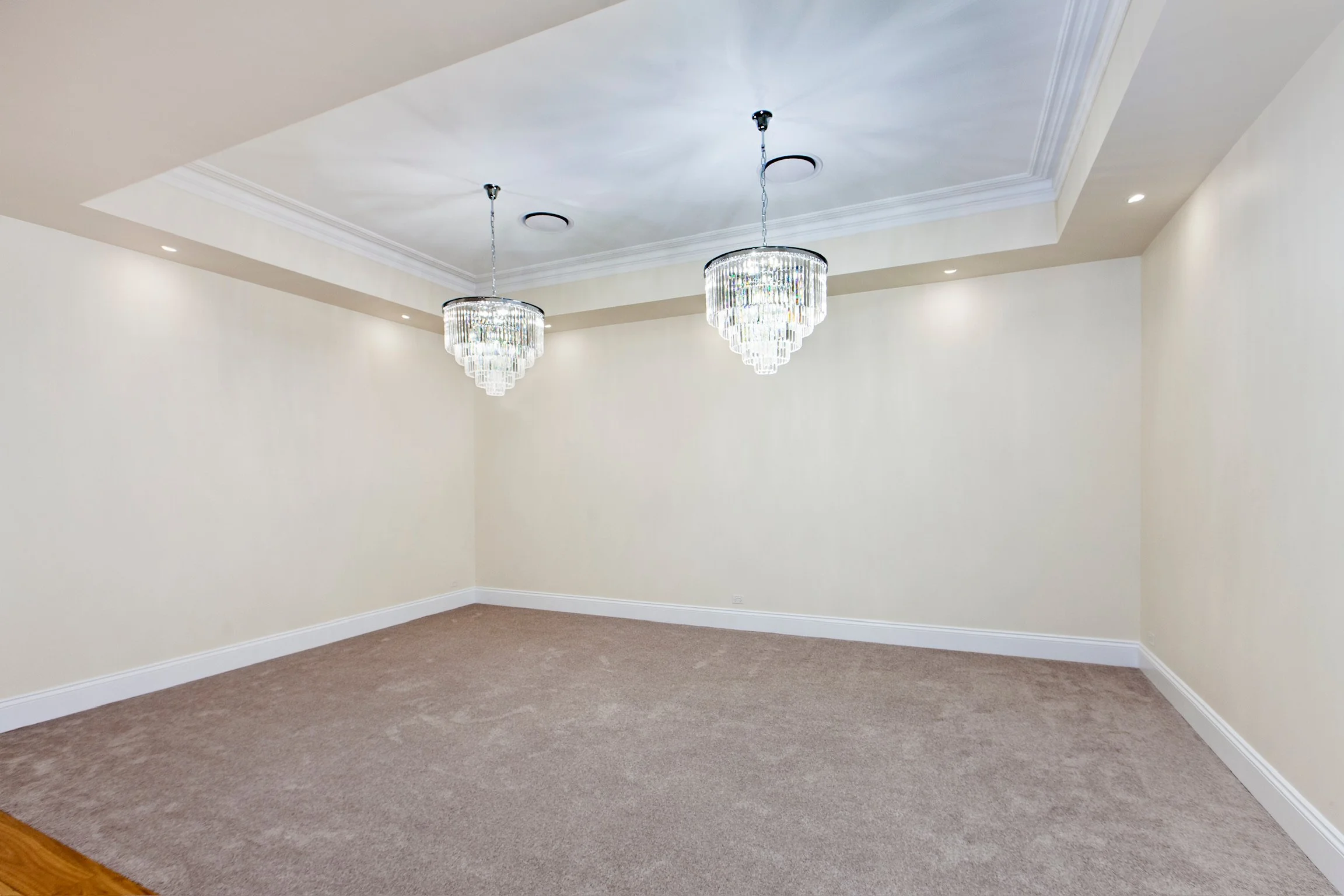 Empty room with beige walls, carpeted floor, white baseboards, and recessed lighting. Two elegant crystal chandeliers are hanging from the ceiling, which has crown molding.