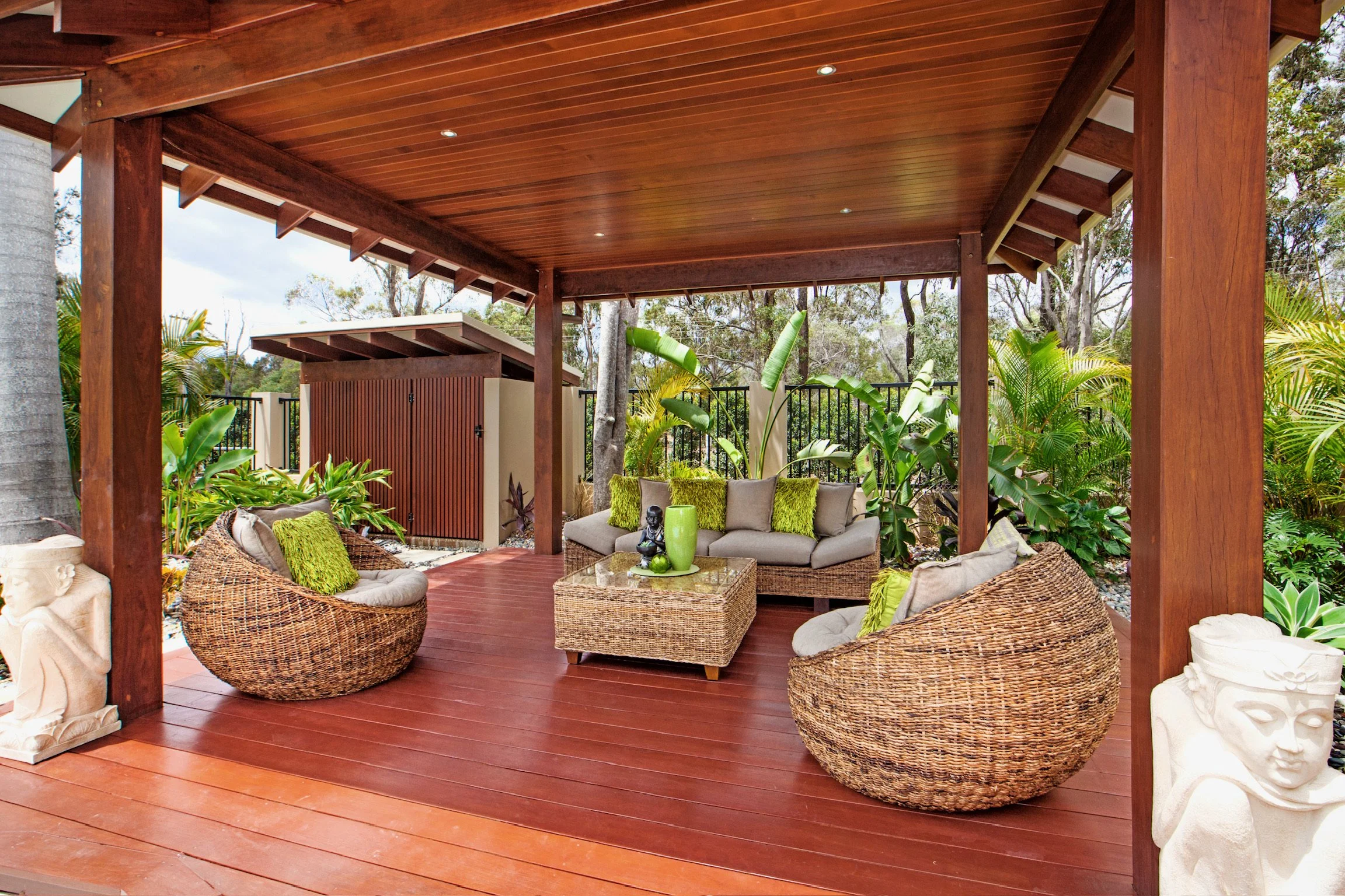 An outdoor patio with wooden flooring and a wooden roof. There are wicker chairs with green pillows and a glass-top coffee table with green vases. Surrounding the patio are lush green plants and trees.