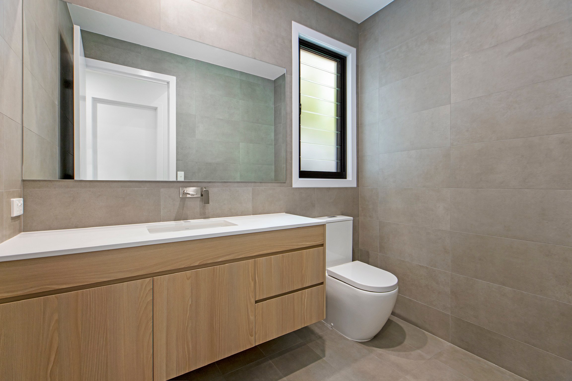 Modern bathroom with large mirror, window, wooden vanity, and toilet.