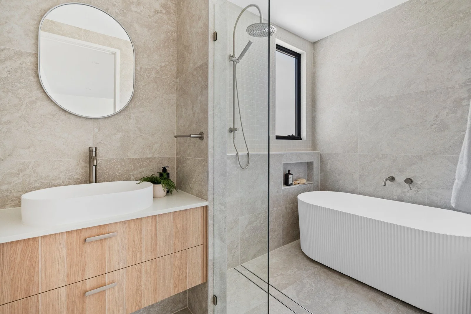 Modern bathroom with a white oval vessel sink and wooden vanity, a round mirror, a walk-in shower with a handheld showerhead, a large bathtub, a window, and neutral beige and gray tiled walls.