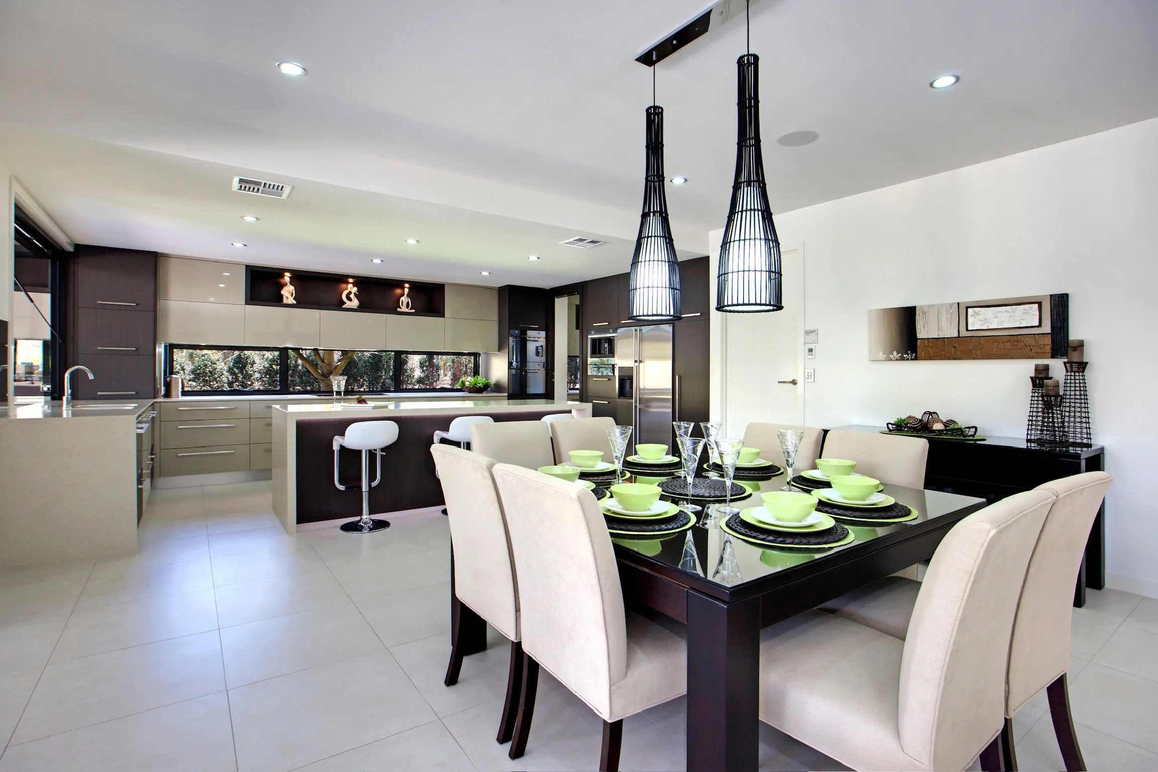 Modern kitchen and dining area with a black dining table set with lime green bowls and glassware, cream upholstered chairs, contemporary black pendant lights, and a kitchen with dark cabinets, stainless steel appliances, and decorative vases on the s