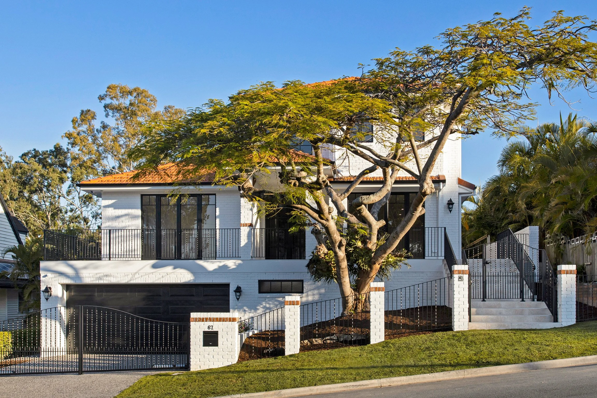 Front view of a modern white brick house with large black-framed windows, a black metal fence, and a mature tree with green leaves in the front yard under a clear blue sky.