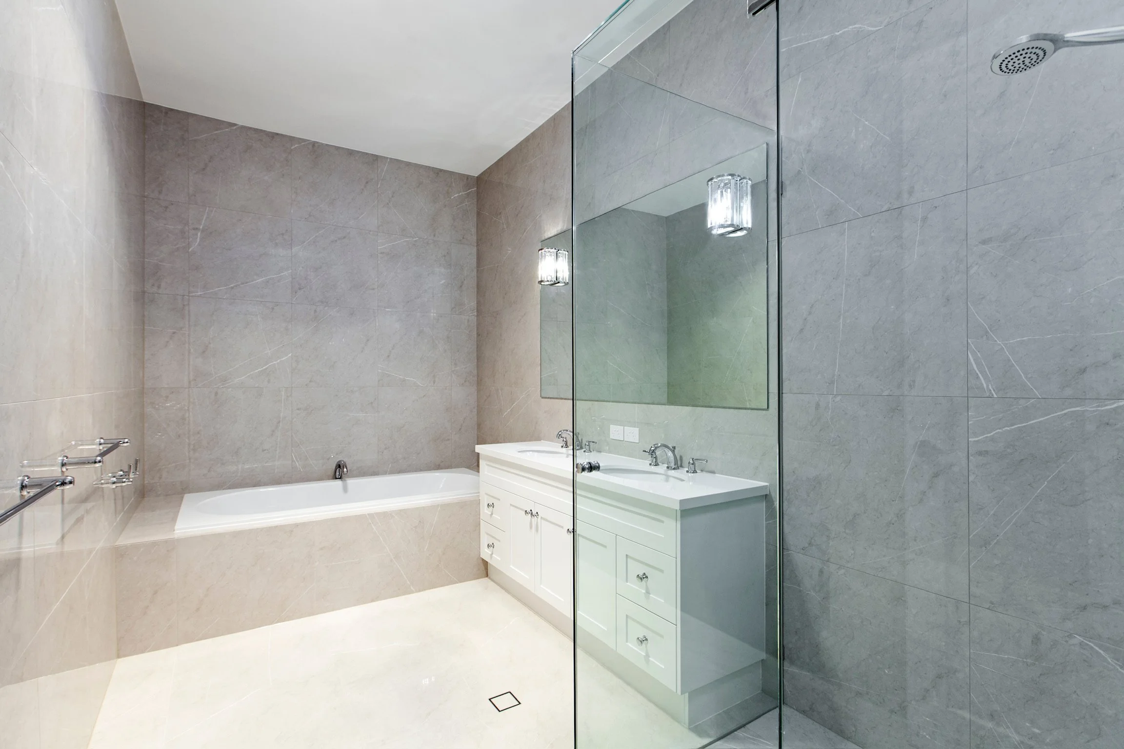 Modern bathroom featuring beige marble walls and floors, a white vanity with a large mirror, two wall-mounted light fixtures, and a glass-enclosed shower area.