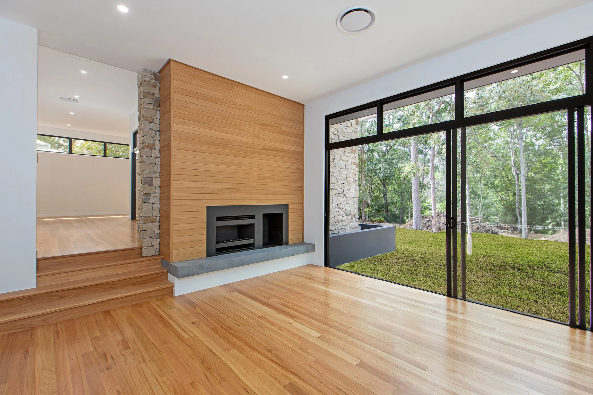 Modern living room with wooden floors, a fireplace, and large glass sliding doors that open to a grassy backyard with trees.