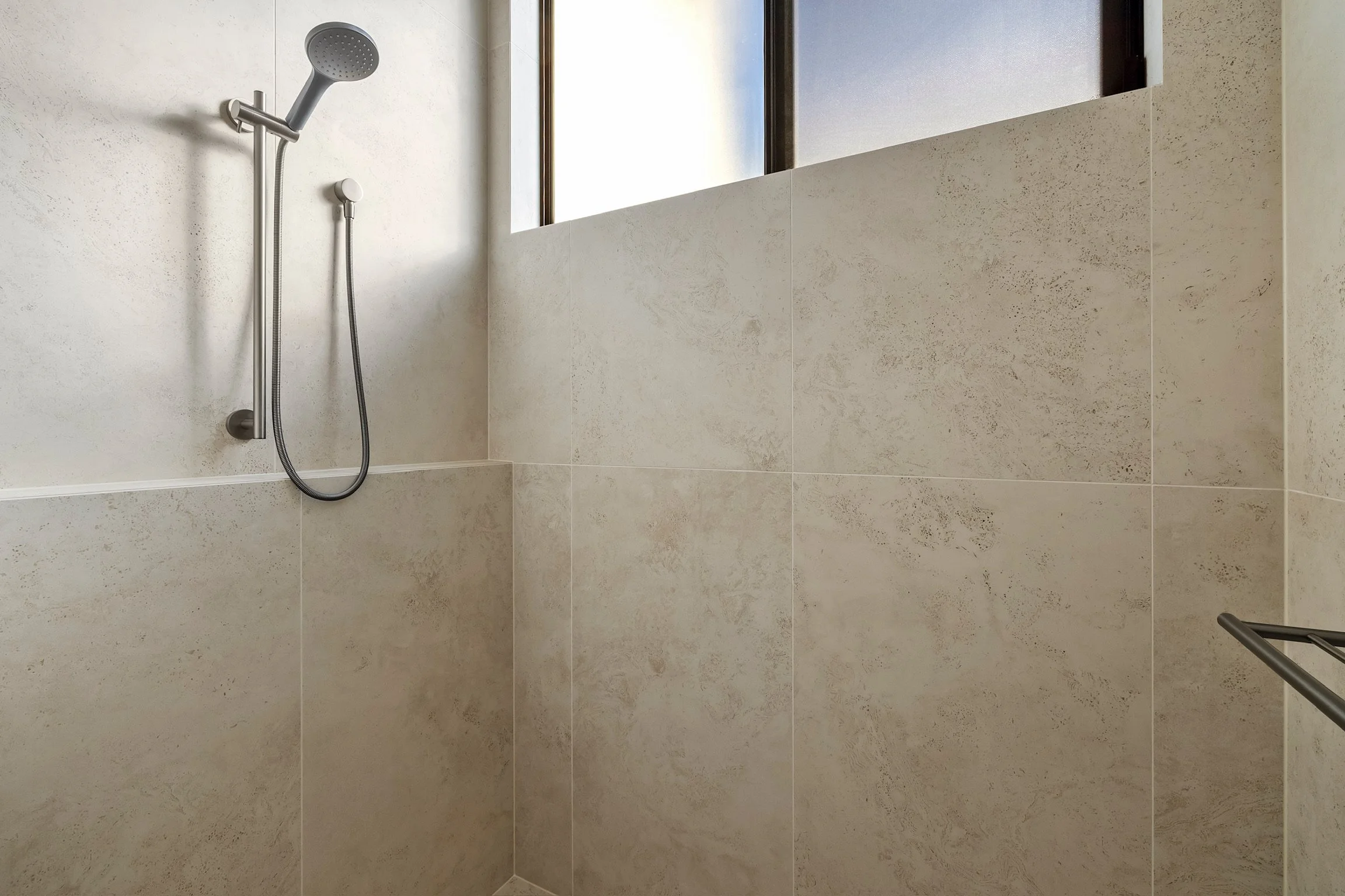 A shower with a handheld showerhead mounted on a wall, next to a window with frosted glass. Beige tiles cover the shower walls.
