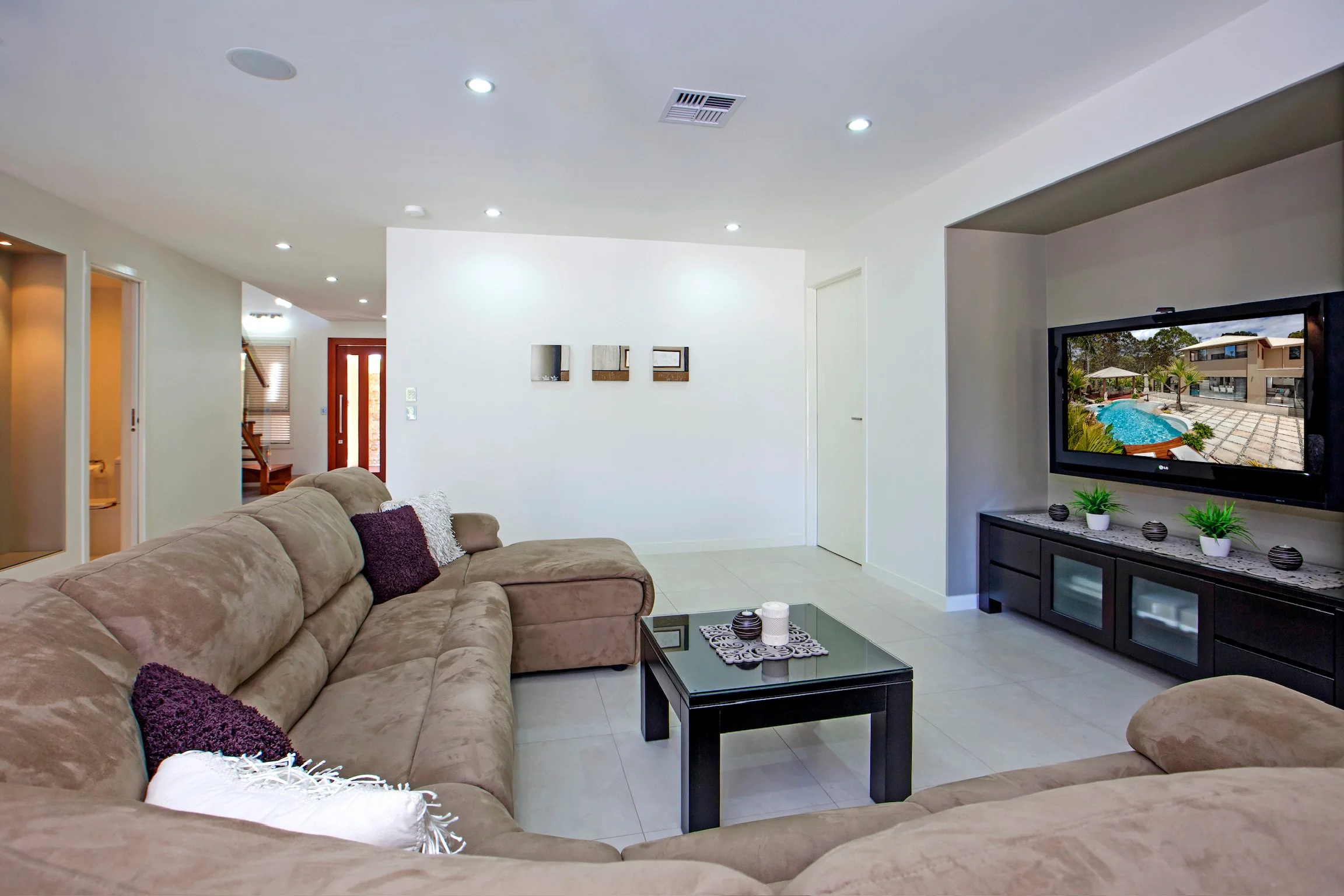 Living room with beige sectional sofa, black coffee table, wall-mounted TV showing a pool scene, white walls, and a doorway with stairs in the background.