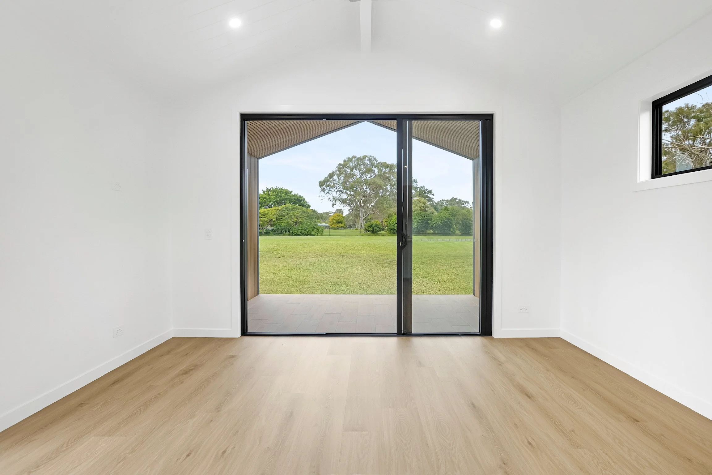 Empty white room with light wooden floors, large sliding glass door opening to a green outdoor field, and a small high window.