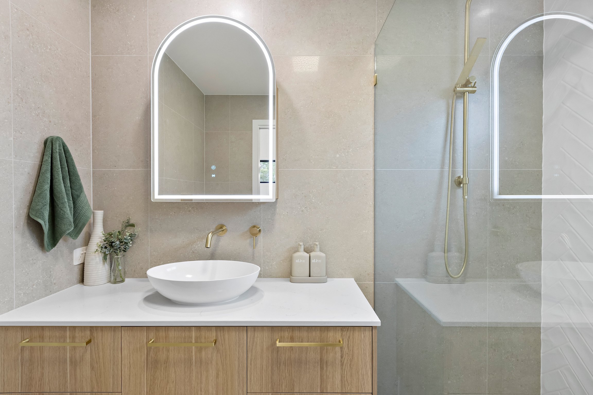 Modern bathroom with beige tiled walls, a wood vanity with gold handles, a white vessel sink, a large mirror with backlit edges, and a shower area with a glass partition, gold fixtures, and a handheld showerhead.