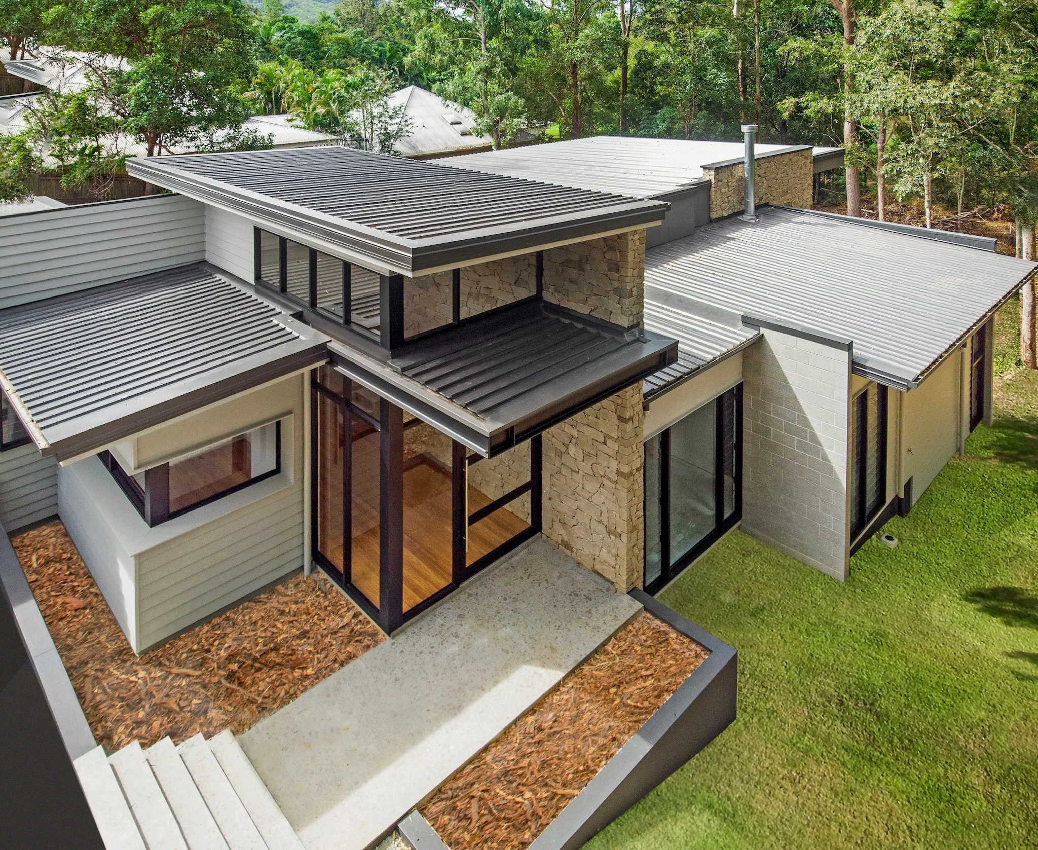 Modern house with metal roofs, large windows, stone accents, and surrounded by trees and green lawn.