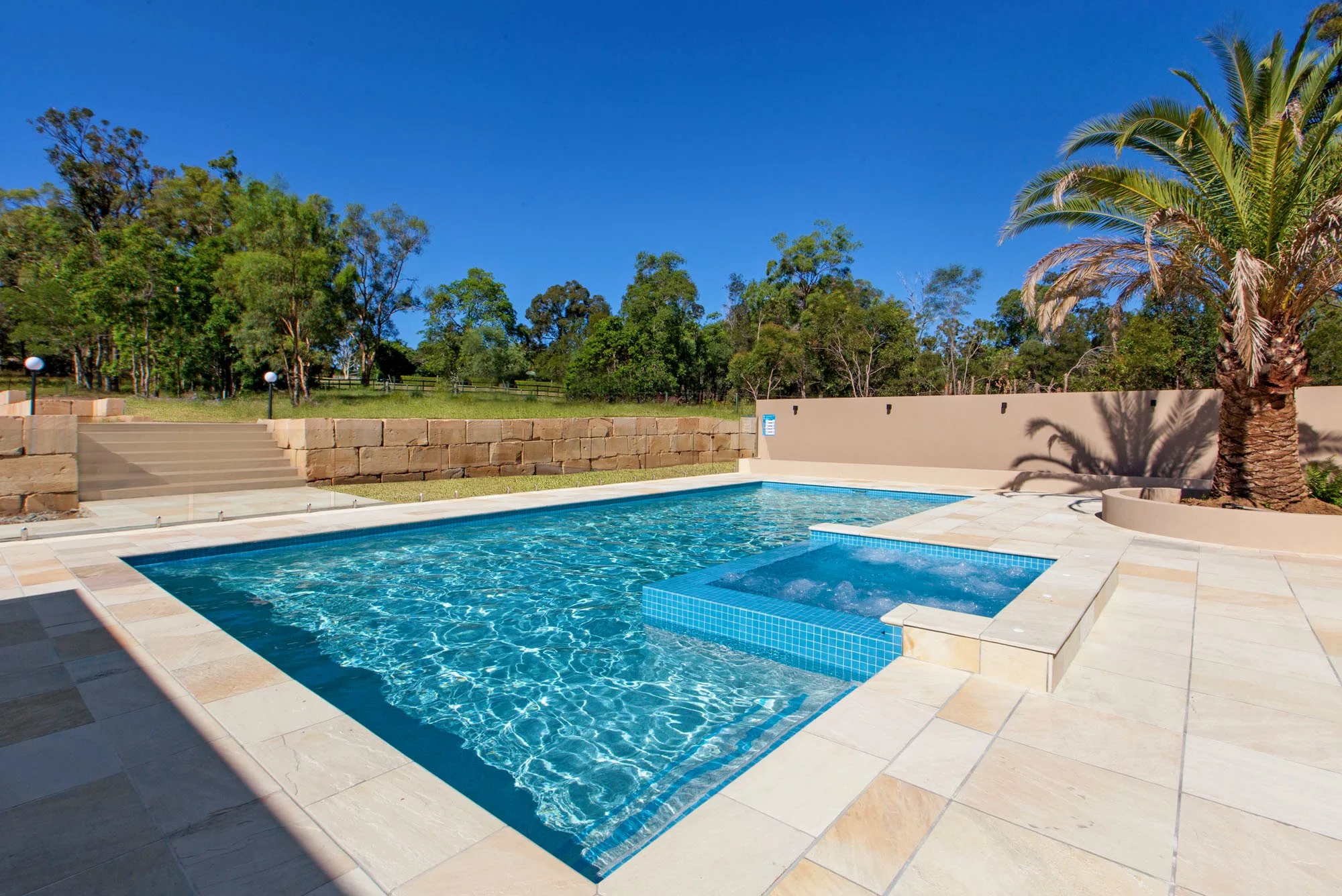 An outdoor swimming pool with a hot tub at one end, surrounded by beige tile decking. There are black lights on the wall, with a white wall and steps leading to a grassy area with trees, including a palm tree on the right. The sky is clear and blue.