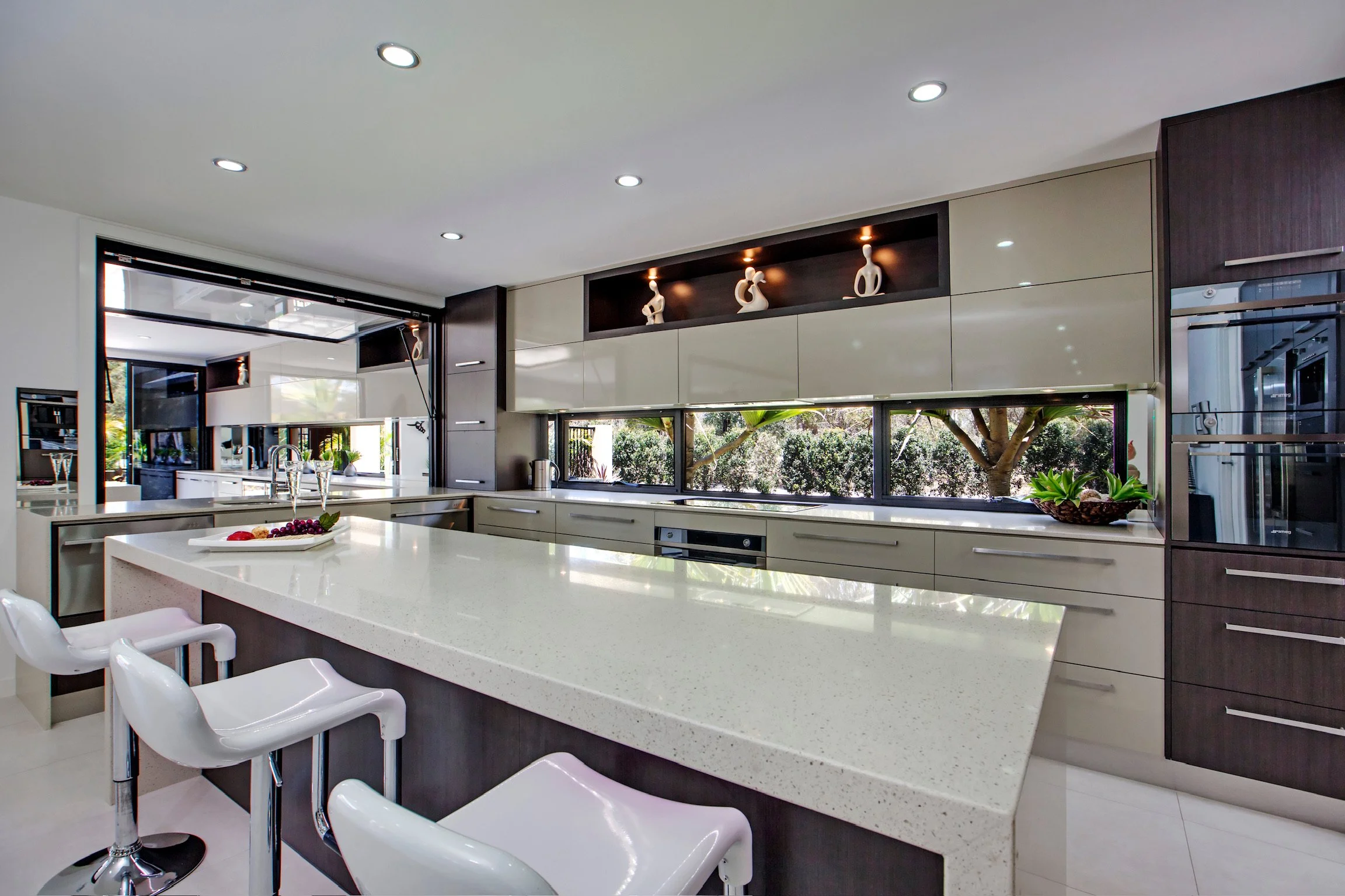 Modern kitchen with white countertops, dark cabinetry, stainless steel appliances, and large windows overlooking greenery.