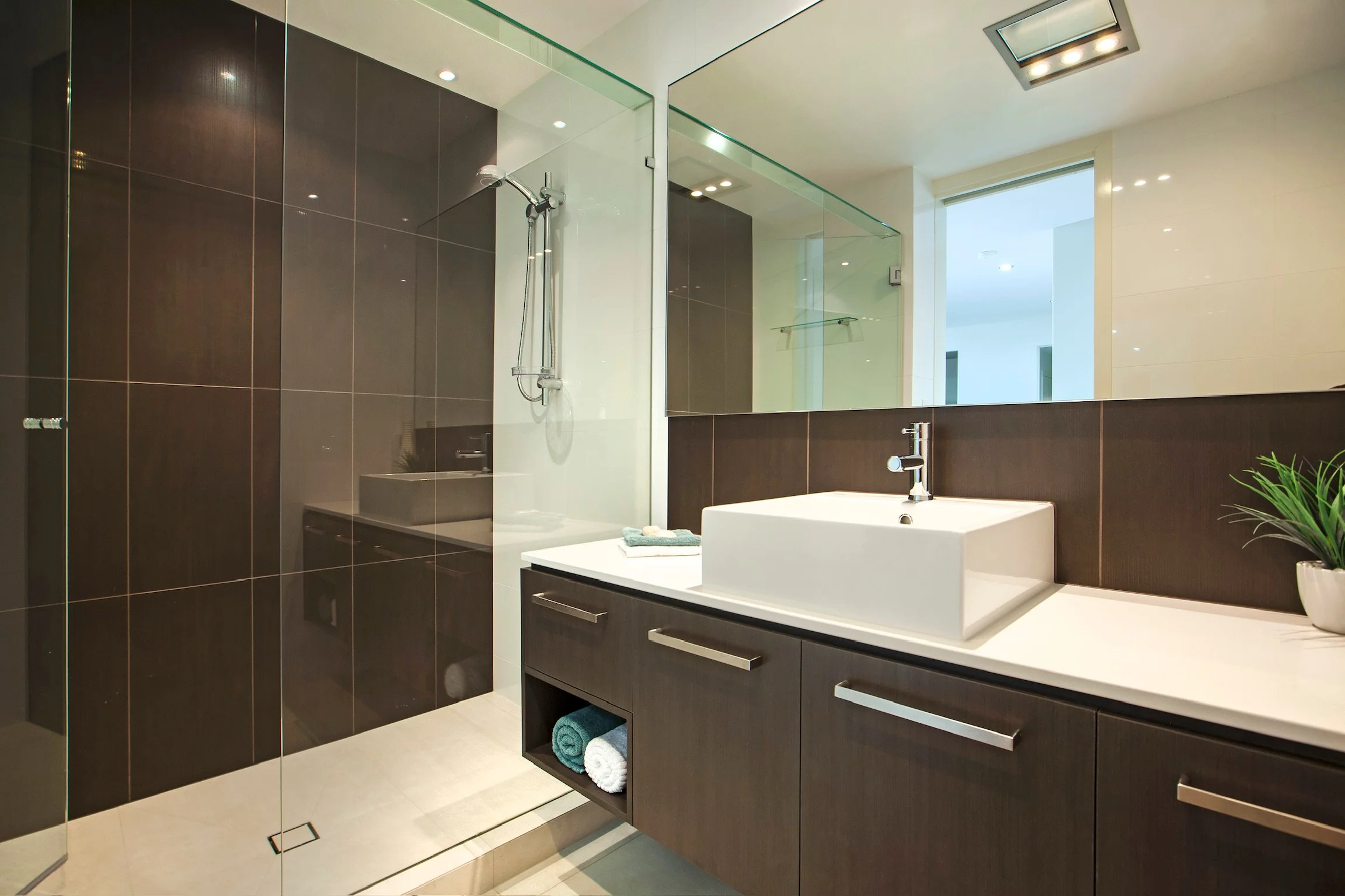 Modern bathroom with dark wood vanity, white countertop, and large mirror. Glass shower enclosure with a mounted showerhead. Light-colored tiles on the floor, a small potted plant on the counter, and a window with natural light.