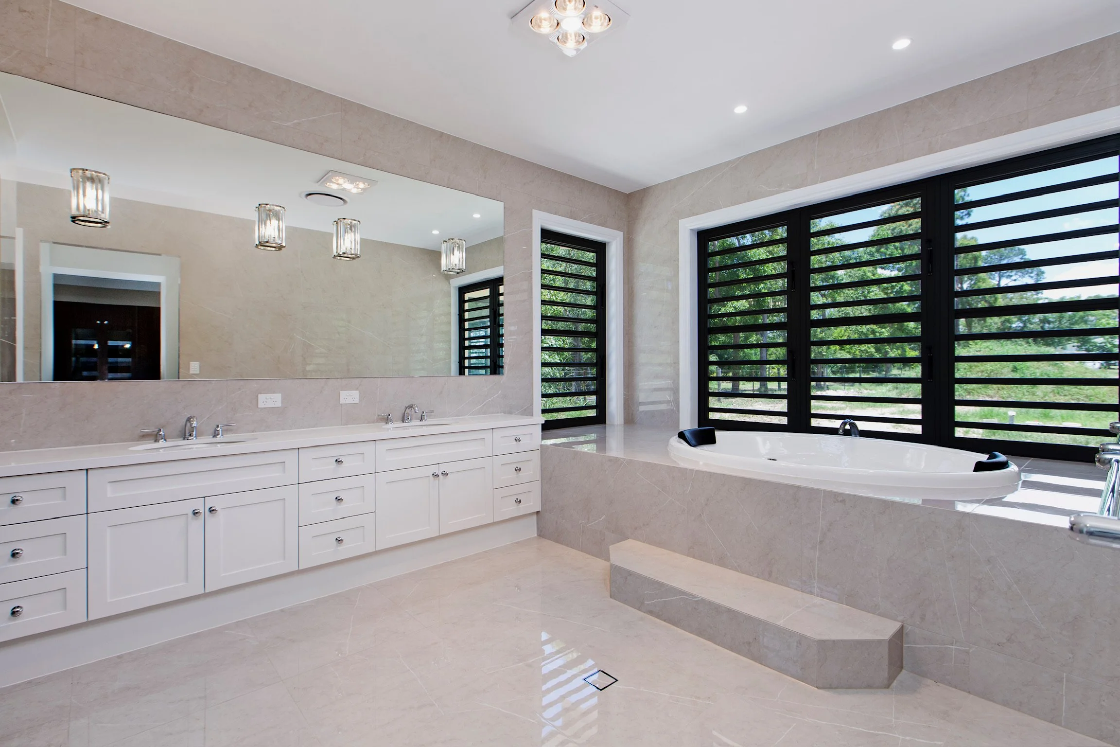 Modern bathroom featuring a large mirror, multiple light fixtures, a double sink vanity with white cabinets, a bathtub with black window shutters, and beige tiled floors and walls