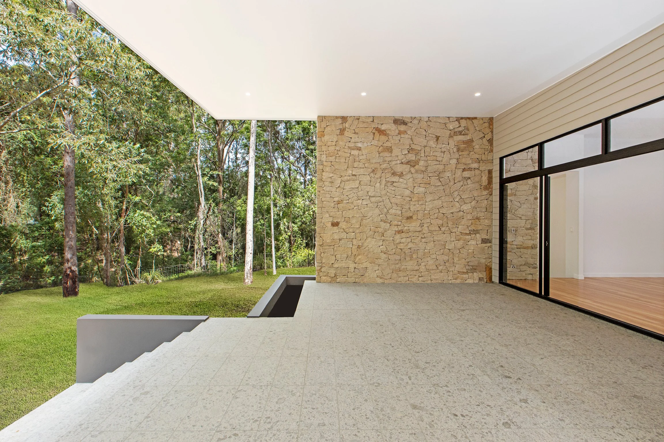 Modern house with a stone wall, large sliding glass doors, and an outdoor patio surrounded by green trees and grass.