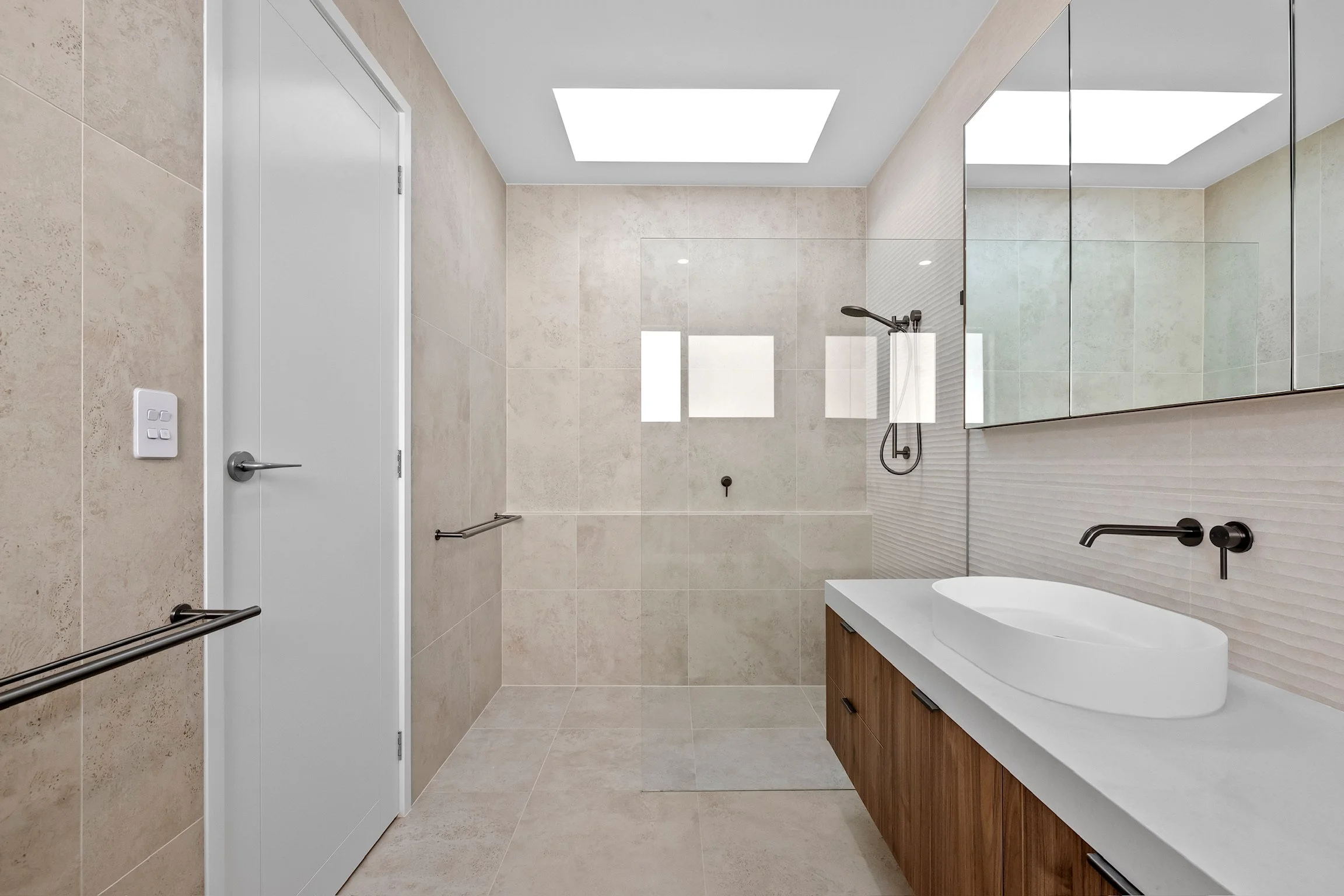 Modern bathroom with beige tiled walls and floor, a large mirror above a wooden vanity with a white sink, a black faucet, and a black showerhead. Skylight windows in the ceiling bring natural light, and there are safety rails on the wall.