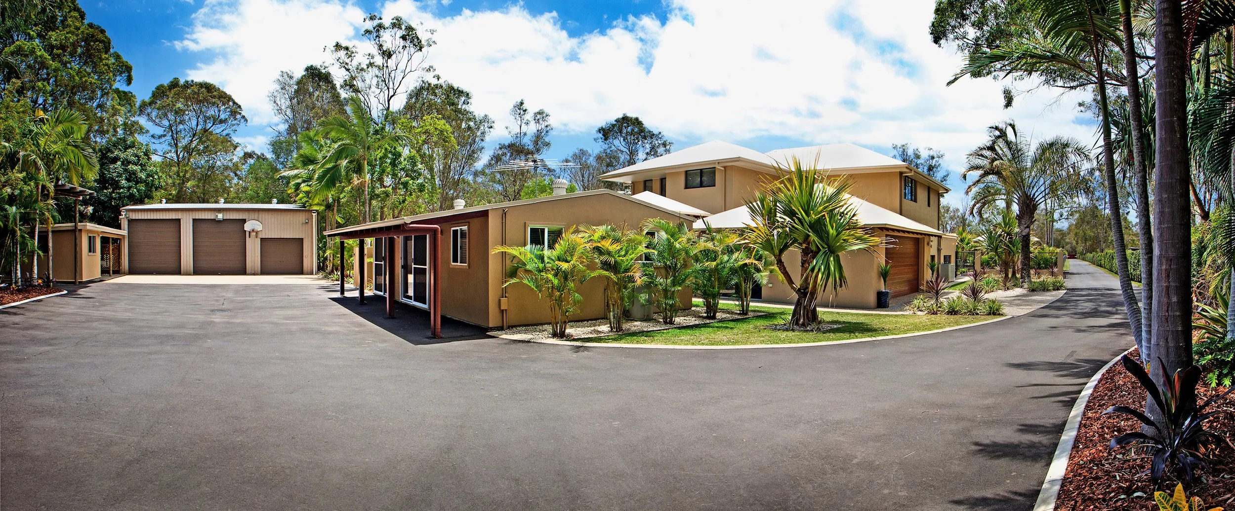 A residential property with multiple buildings, a large paved driveway, lush green tropical plants, and palm trees, under a partly cloudy sky.