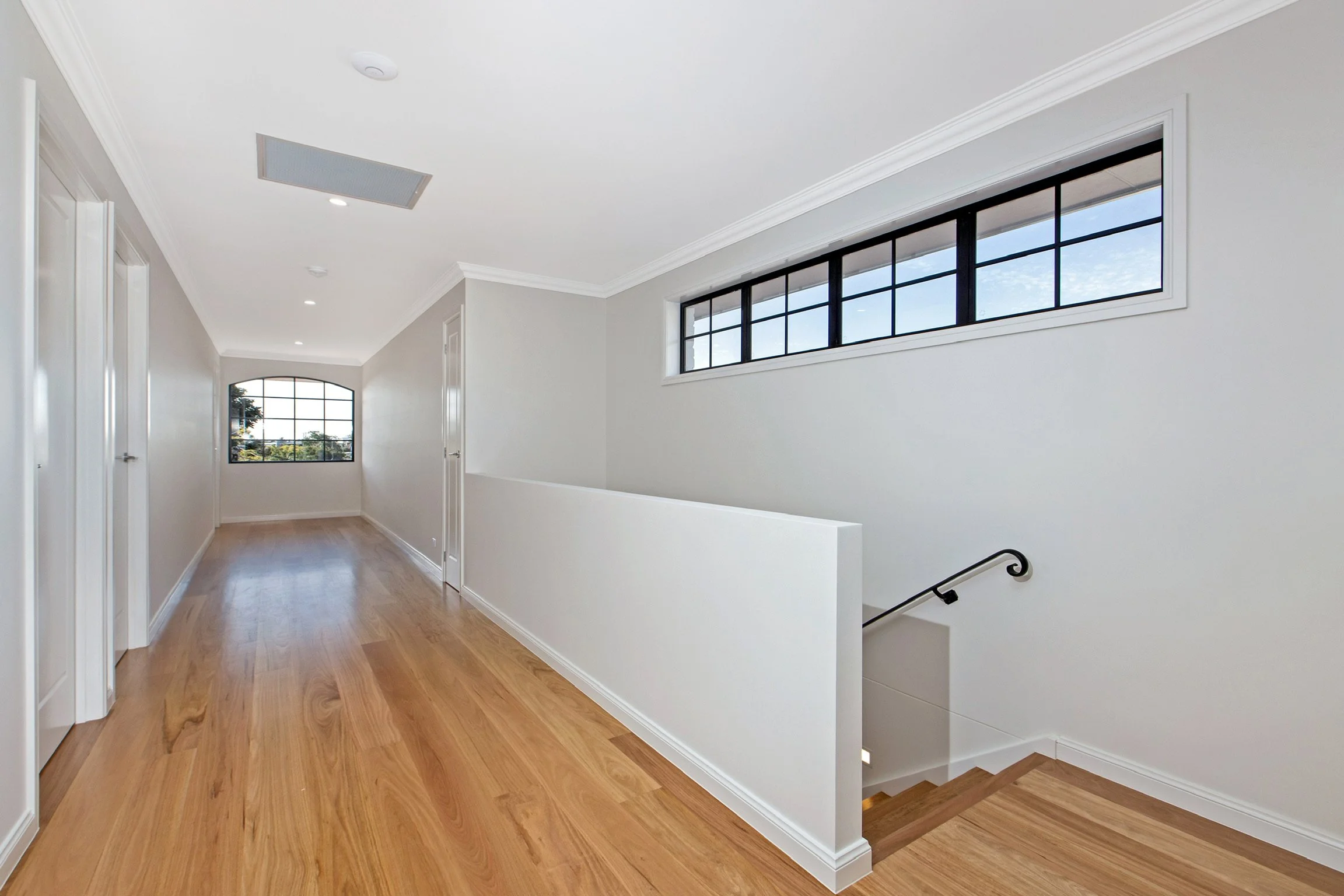 A bright, modern interior hallway with white walls, wooden flooring, rectangular windows along the ceiling, and a staircase with a black handrail.