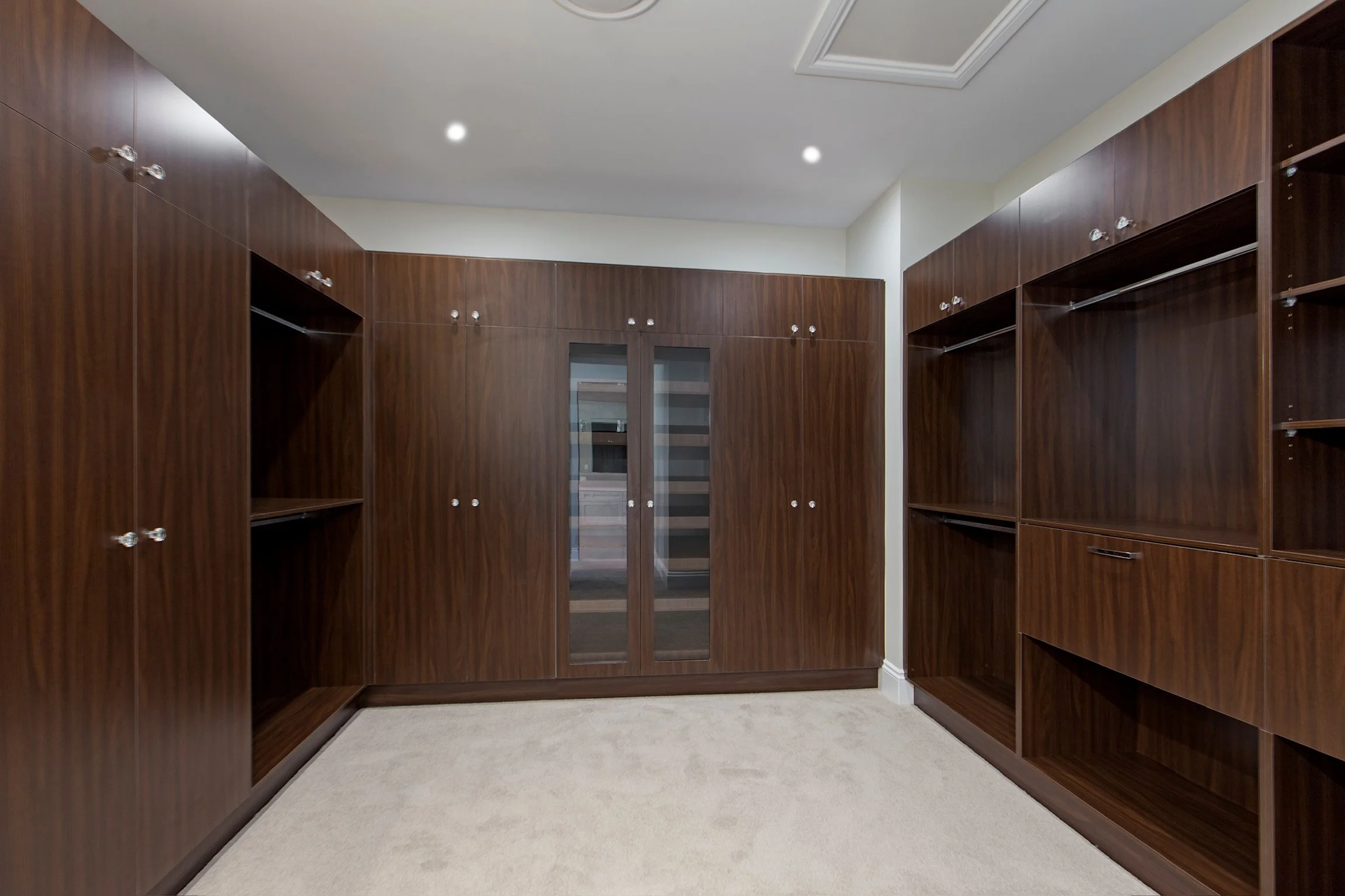 Empty walk-in closet with dark wood cabinets and shelves, beige carpet, and a white ceiling with integrated lighting.