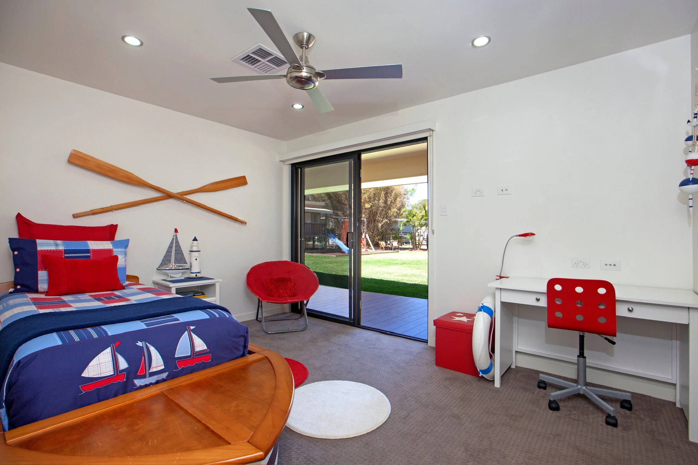 A bedroom with nautical themed decor. It has a bed with a red, blue, and white sailboat bedspread, a wooden bed frame, and pillows. There is a small white bedside table with a model sailboat and lighthouse, and a red round chair. A ceiling fan and re