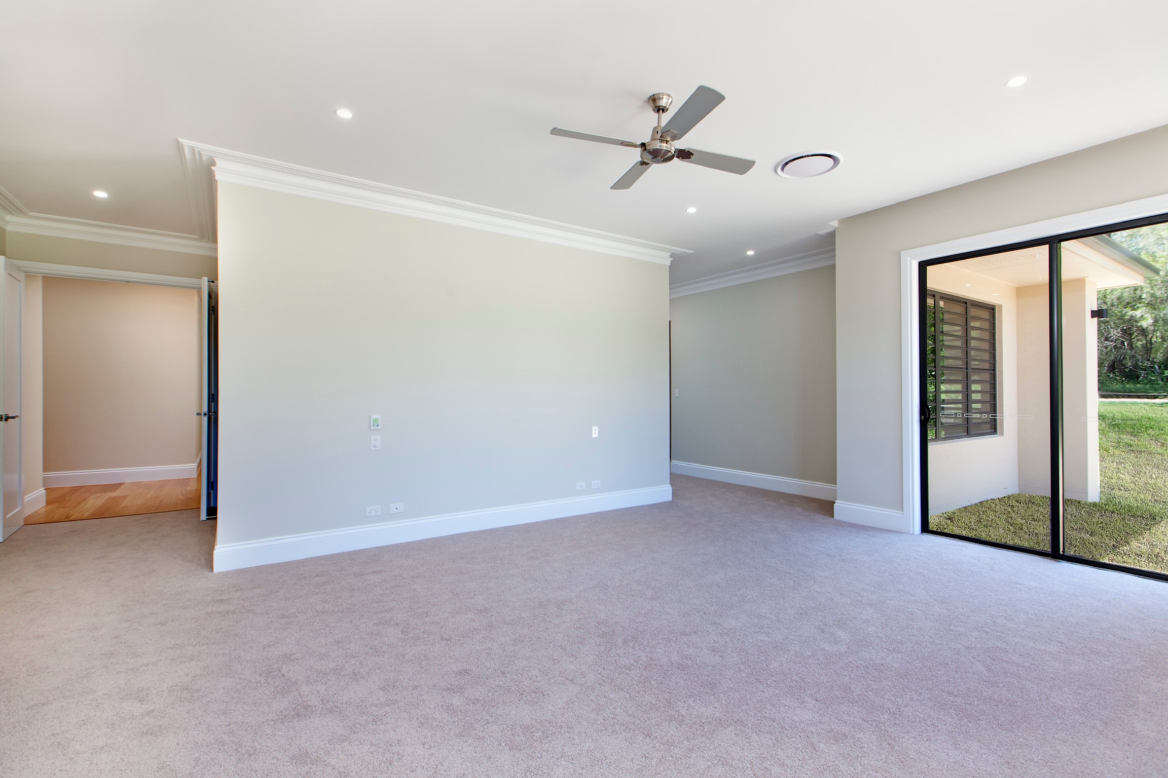 Empty living room with beige carpet, off-white walls, ceiling fan, and sliding glass door leading outside to yard.