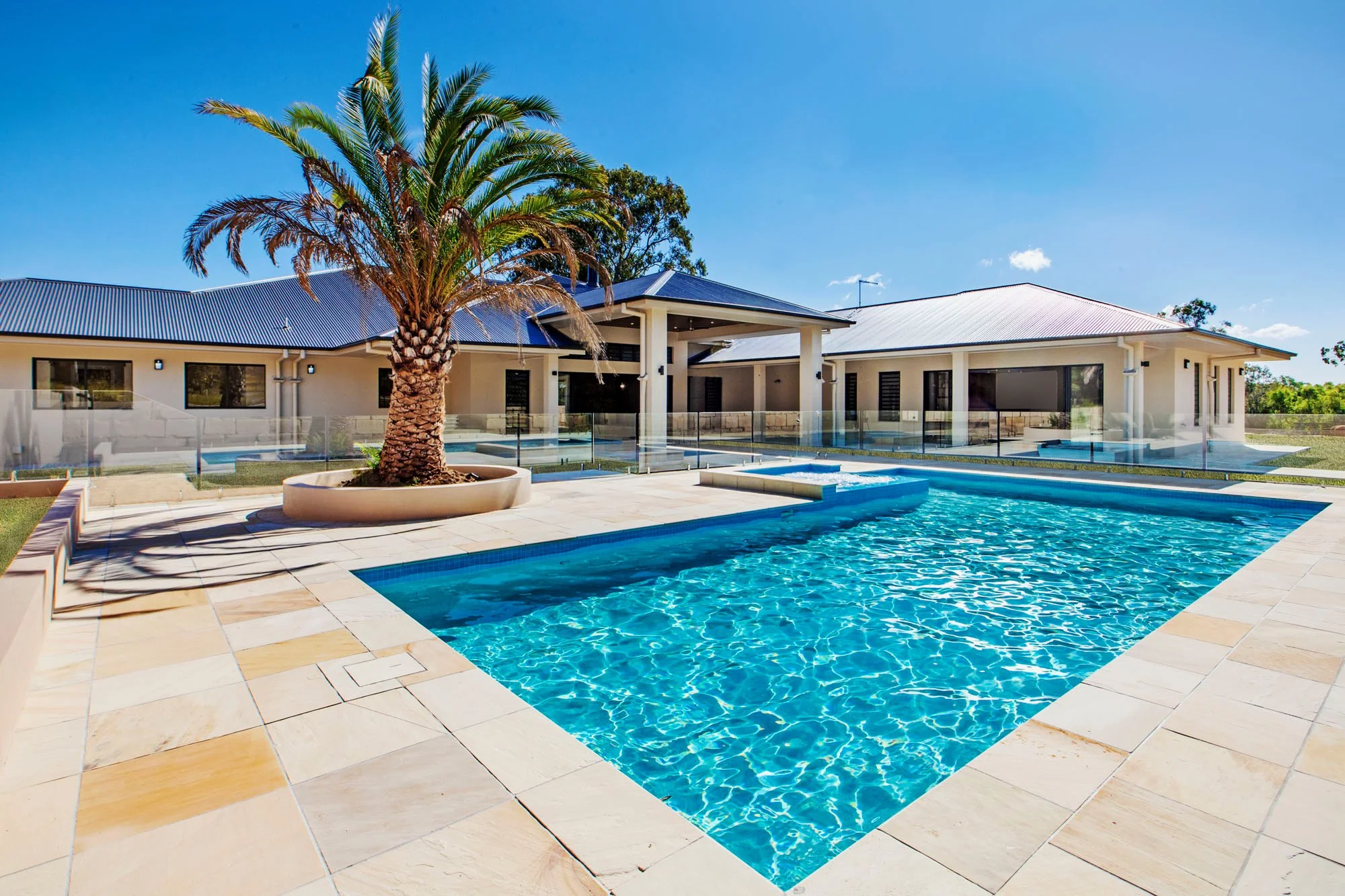 A modern house with a pool in the backyard, featuring a palm tree, a clear blue sky, and glass fencing around the pool area.