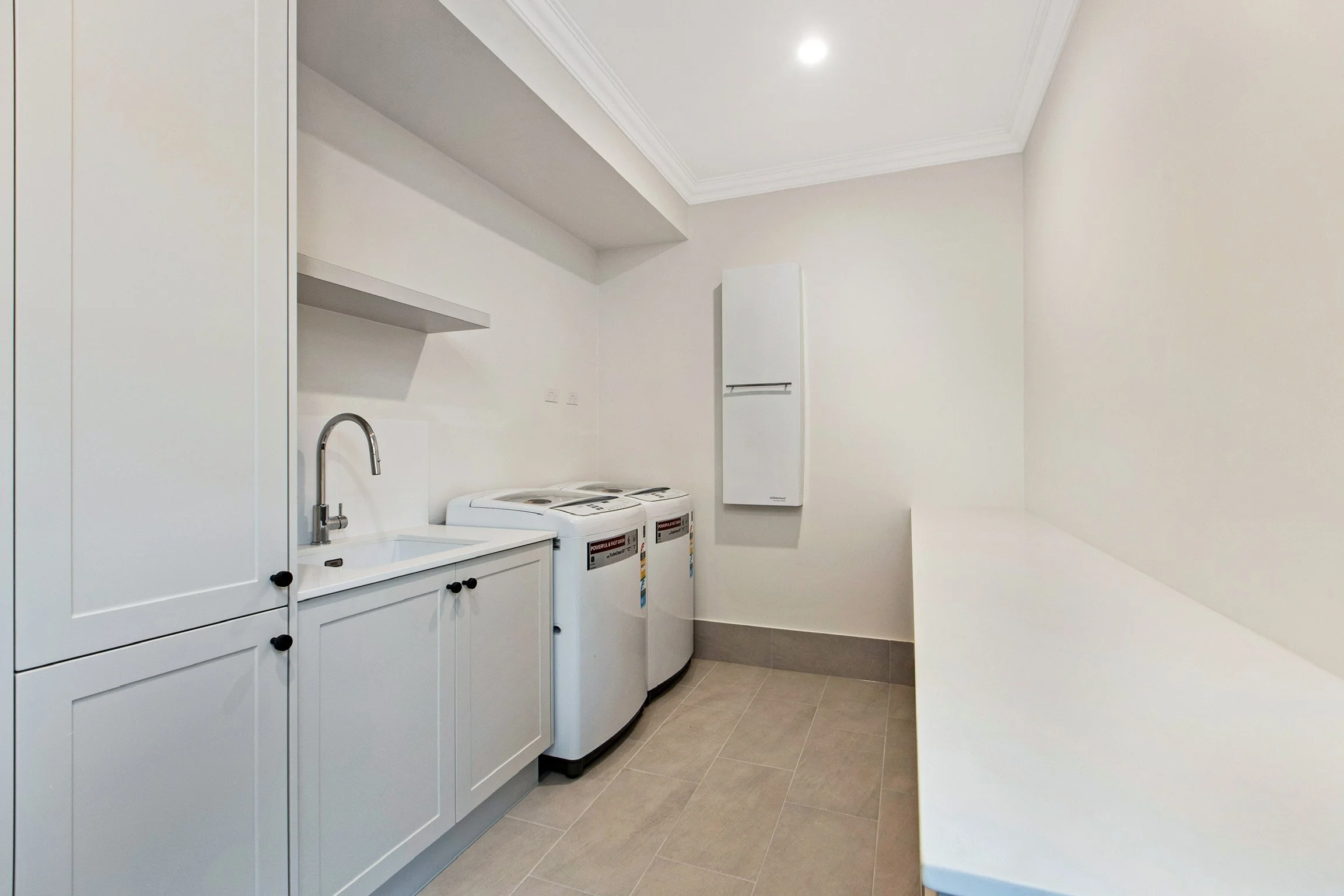 A laundry room with white cabinets, a sink with a faucet, two top-loading washing machines, white walls, and beige tiled flooring.