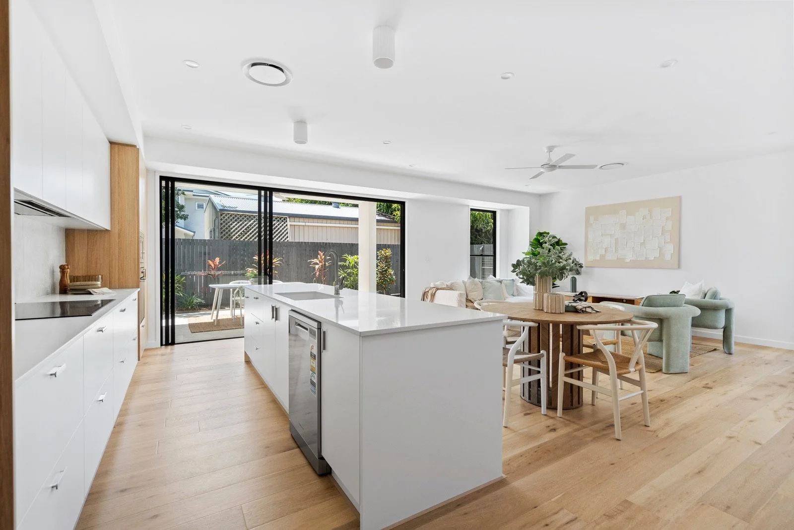 Modern open-plan kitchen and living area with white cabinets, a kitchen island, and a round dining table with chairs, overlooking a backyard with outdoor furniture.