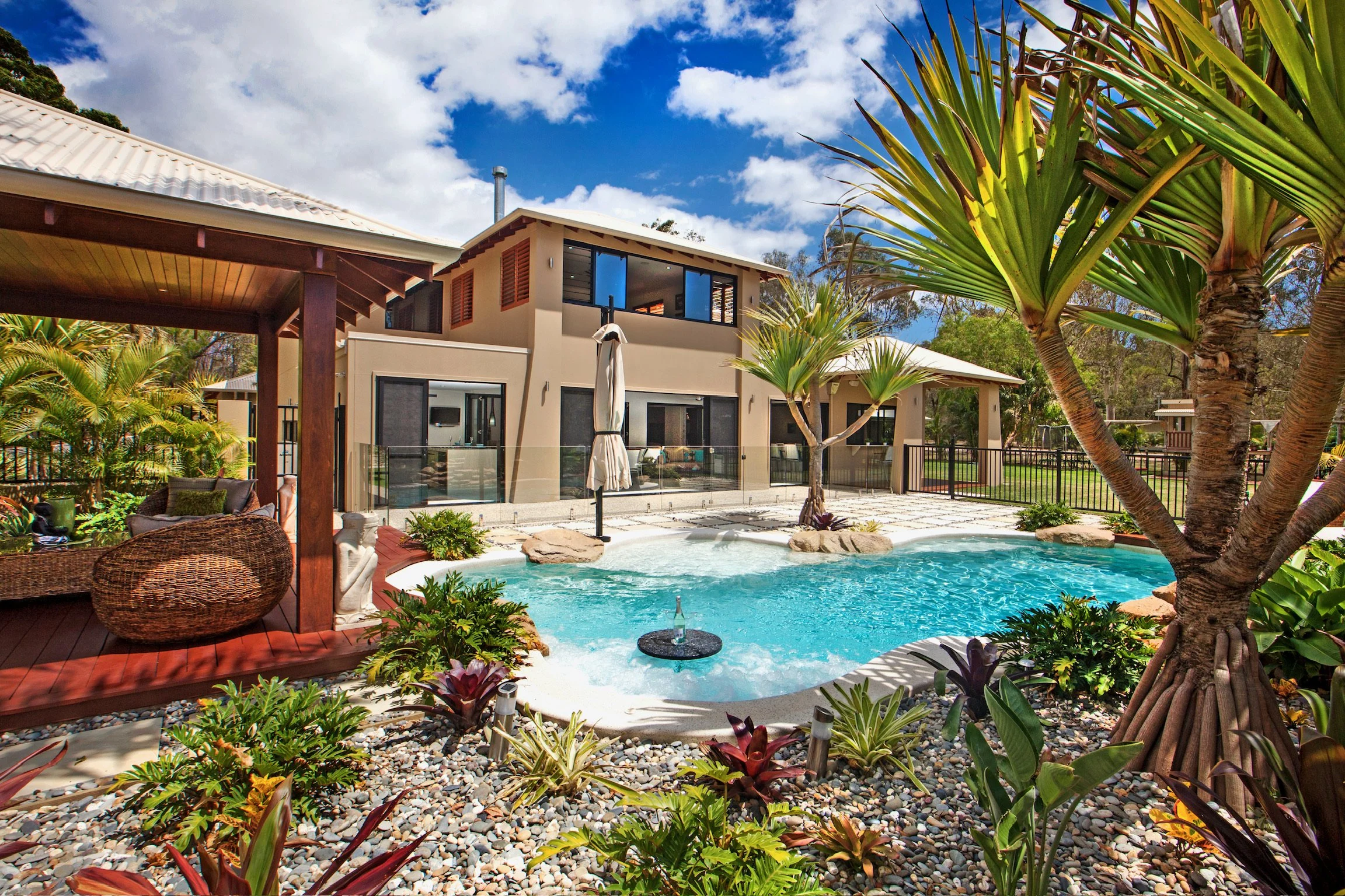A backyard with a swimming pool, surrounded by tropical plants and trees, with a modern two-story house in the background under a partly cloudy sky.