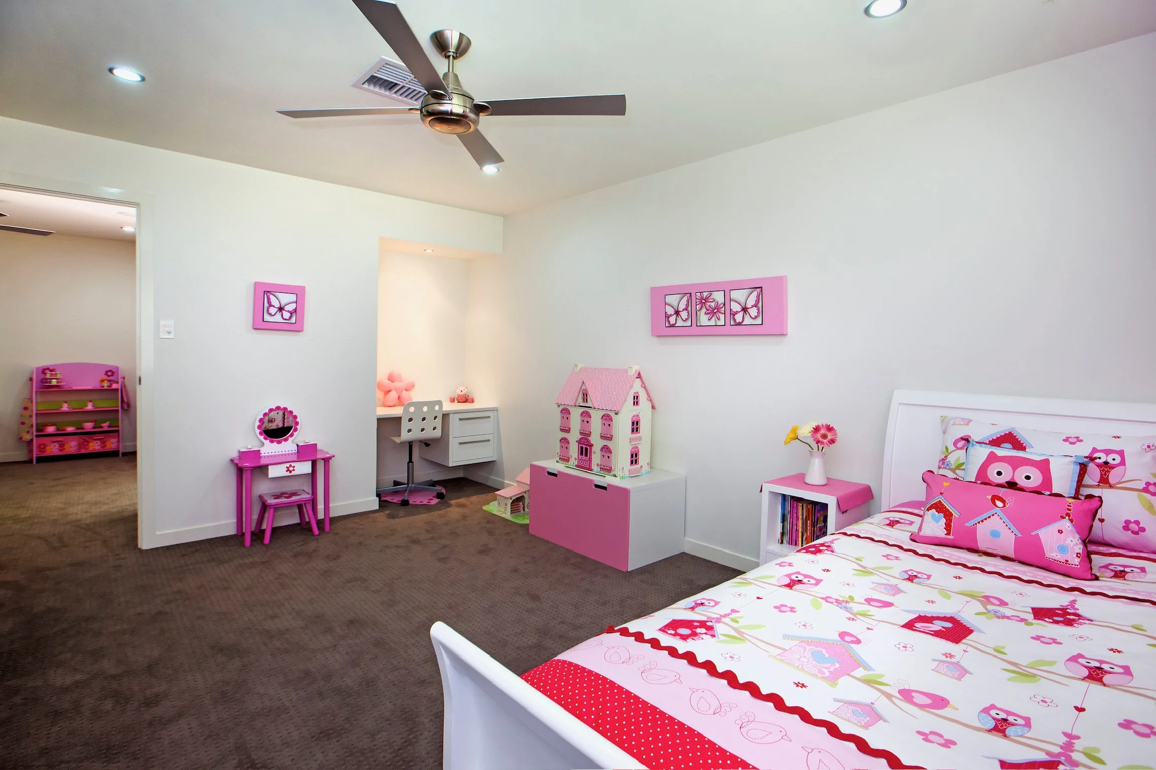 A children's bedroom with a bed featuring pink, red, and white owl-themed bedding, pink accents, and pink wall art with butterflies and flowers. The room has a white nightstand with books, a pink dollhouse, a small pink vanity with a mirror, a pink c