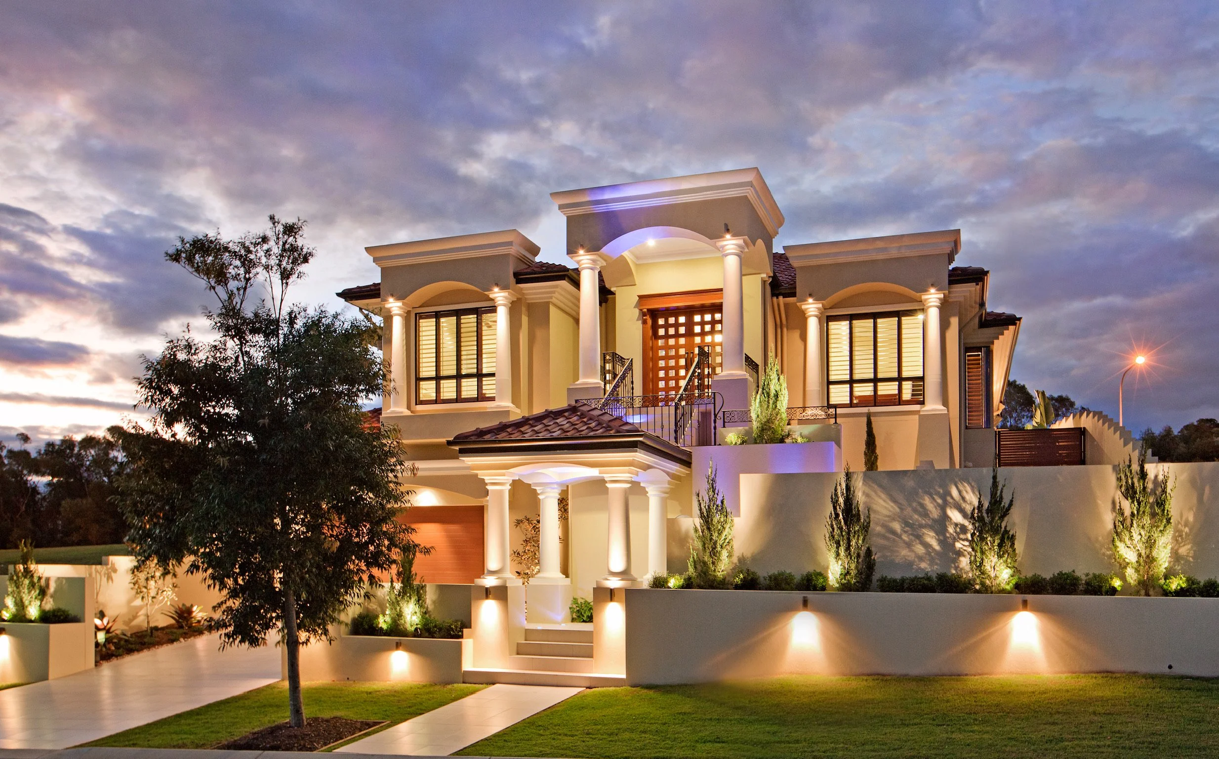 A luxurious modern two-story house illuminated at dusk, with tall columns at the entrance, large windows with shutters, and landscaped front yard with trees and lighting.