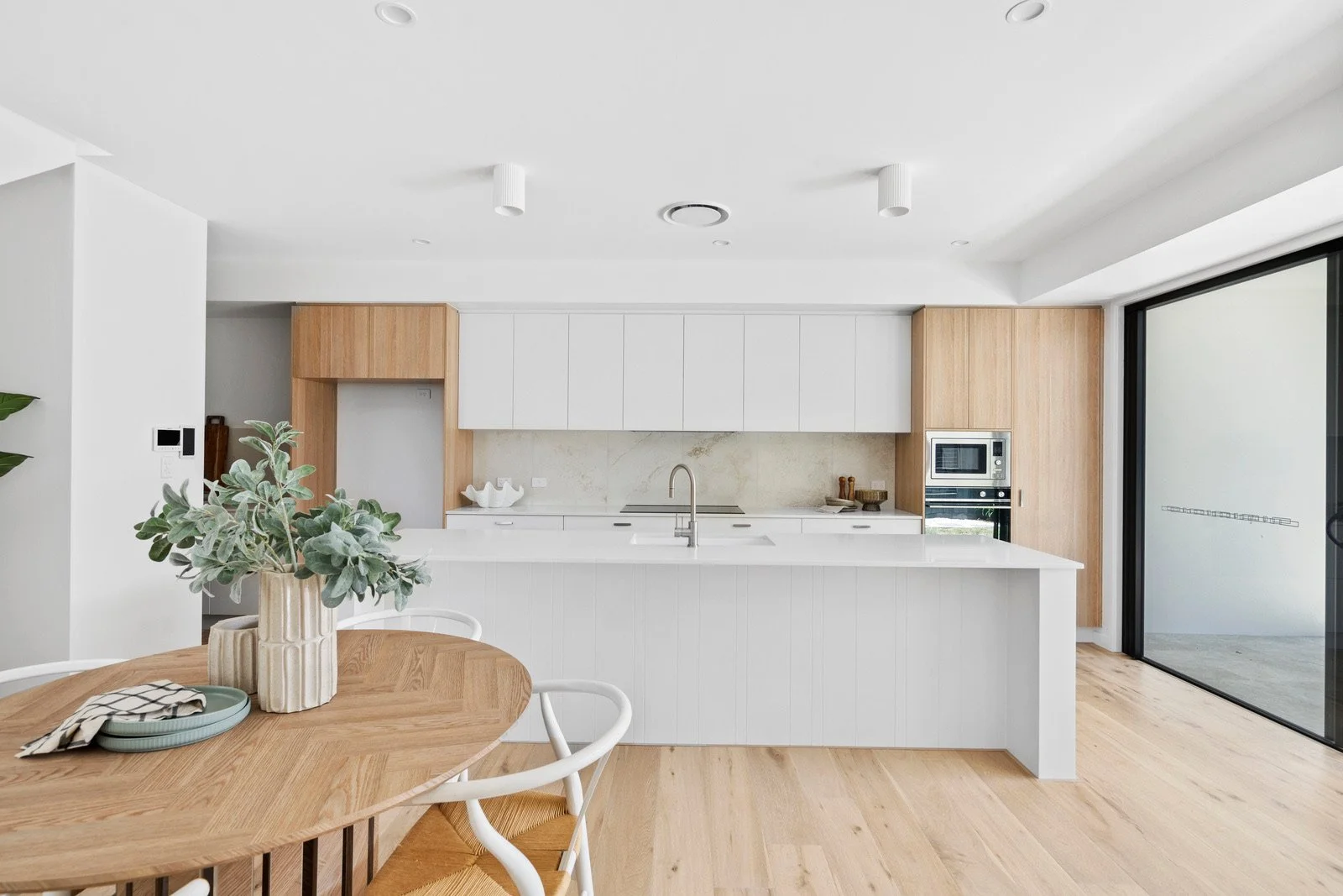 Modern kitchen with white cabinetry, wood accents, and a large sliding glass door, featuring a dining table with a potted plant centerpiece.