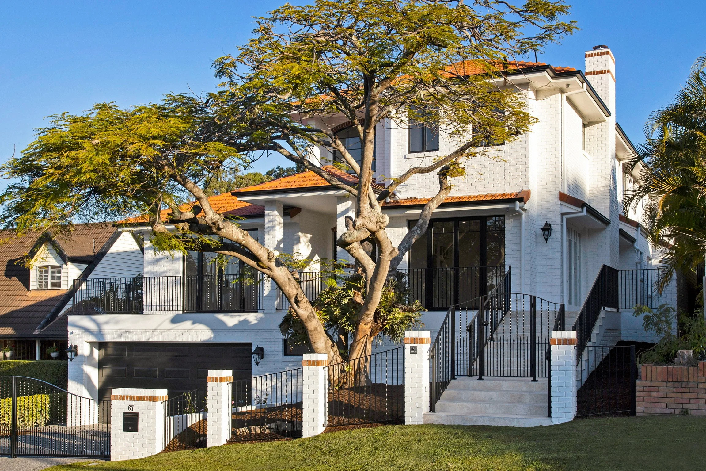 Modern white brick house with a red tile roof, large windows, and a black front door. A large tree with green leaves is in front of the house, and there is a black metal fence and stairs leading up to the entrance. The sky is clear and blue.