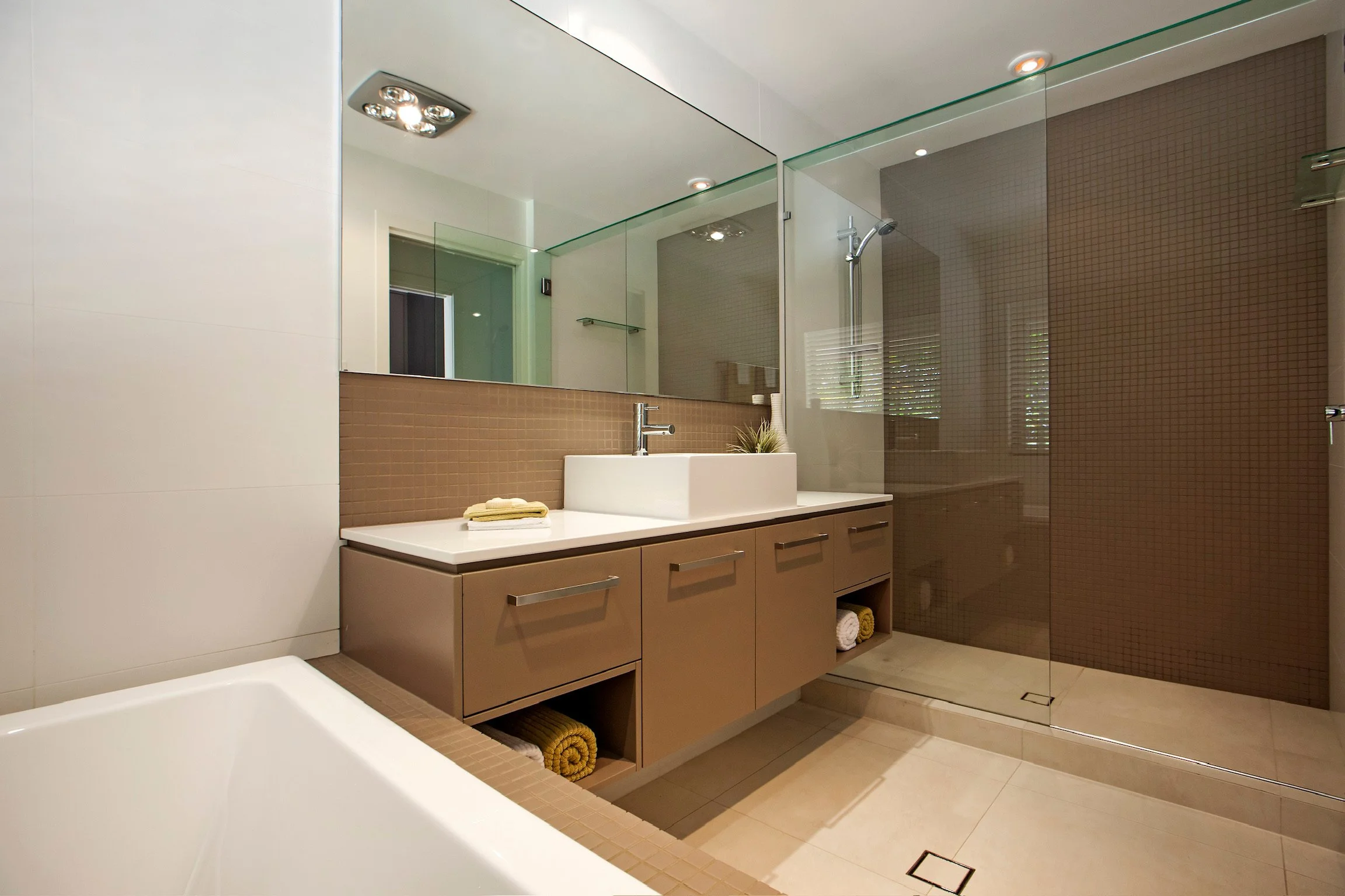 Modern bathroom with beige and brown tiles, vanity with a rectangular sink, mirror above, glass shower enclosure, and rolled towels.