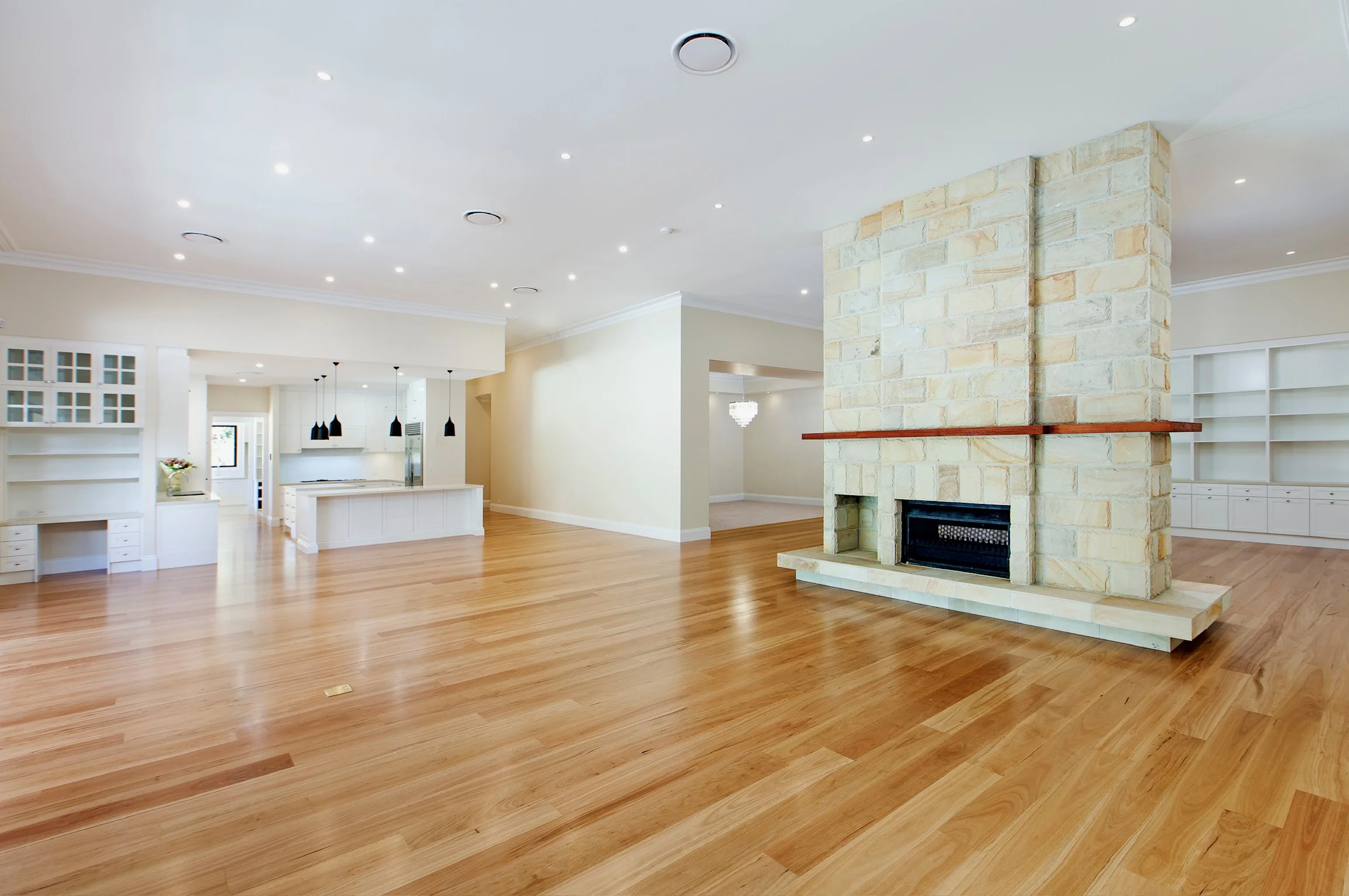Spacious living area with hardwood floors, a stone fireplace, white walls, built-in shelves, and modern lighting fixtures.