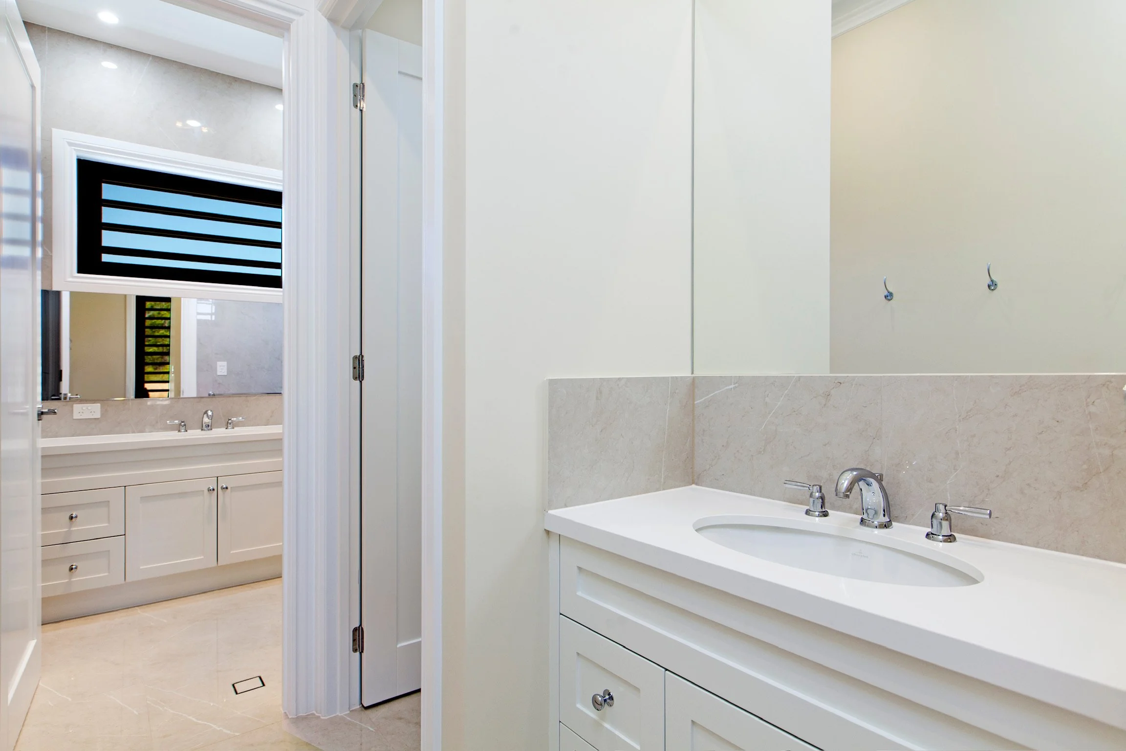 A clean modern bathroom with a white vanity and sink, beige marble tile, a large mirror, and a window with black horizontal blinds.