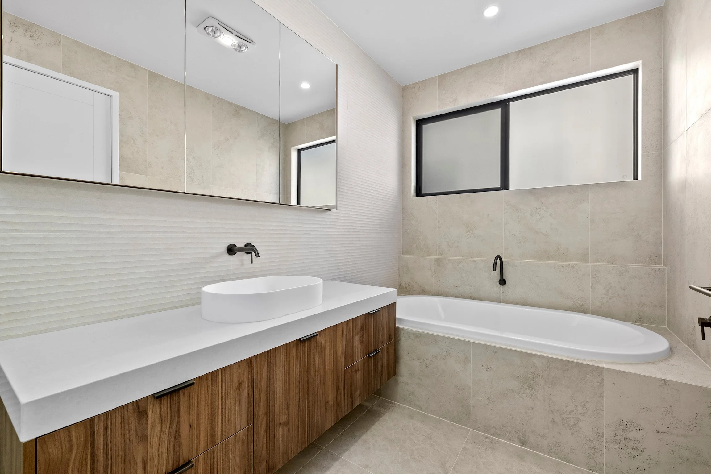 Modern bathroom with beige tiled walls, a large mirror, a white countertop with a vessel sink, and a wooden vanity cabinet. There is a bathtub with a black faucet and a high horizontal window with frosted glass.