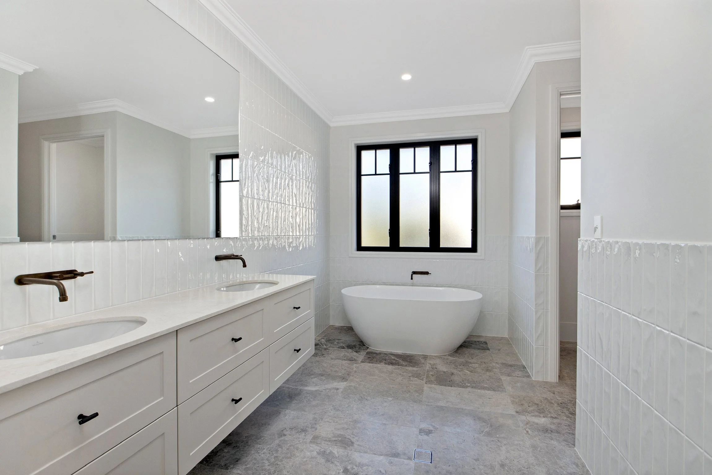 Modern bathroom with dual sinks, large mirror, white cabinetry, freestanding bathtub, black framed window, neutral wall tiles, and gray floor tiles.