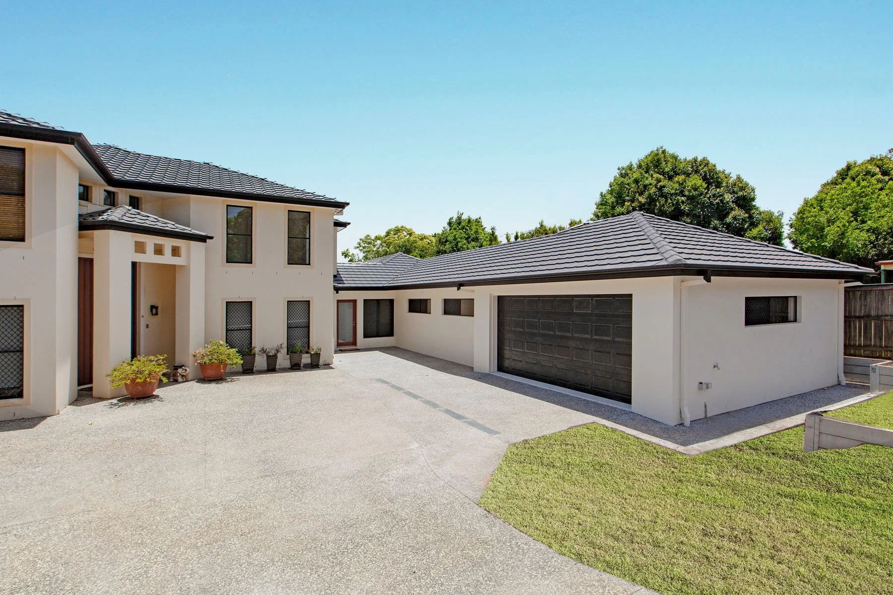 Modern two-story house with white exterior walls, black roof tiles, a paved driveway, and a detached garage, surrounded by green trees and grass.
