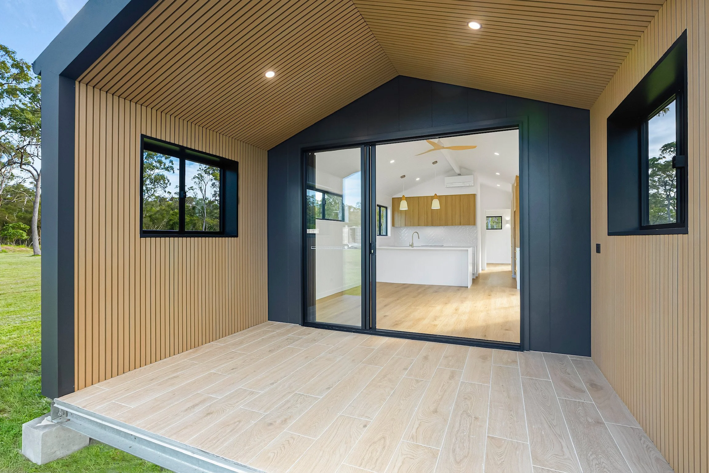 View of a modern outdoor patio with wood slat ceiling and walls, large sliding glass doors leading to an interior living space, and surrounding greenery.
