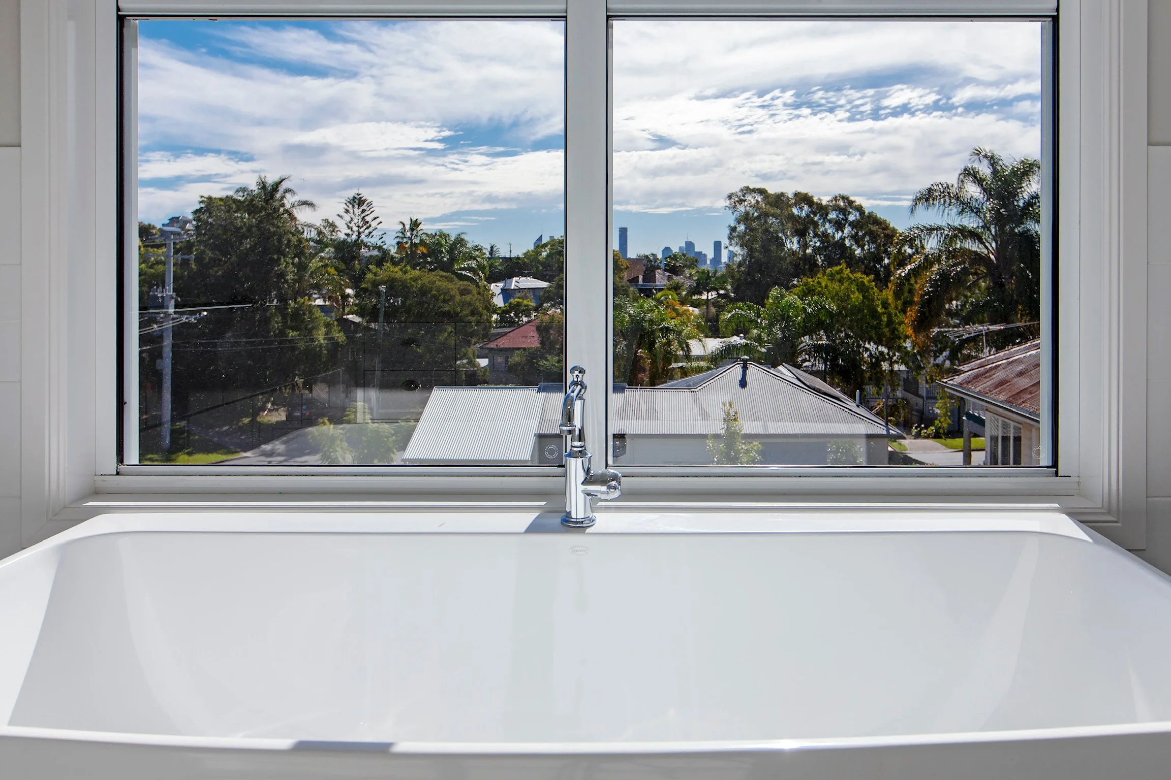 View through a window showing a neighborhood with houses, trees, and a distant city skyline under a partly cloudy sky.