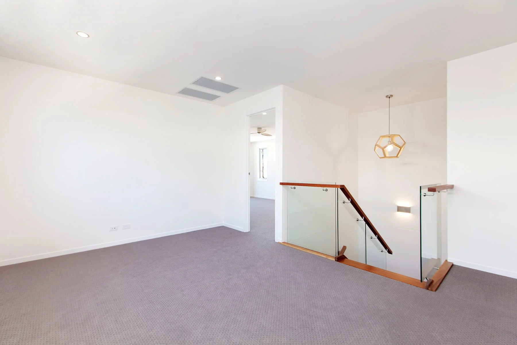 Empty modern living space with white walls, gray carpet, staircase with glass railing, and minimalist light fixtures.